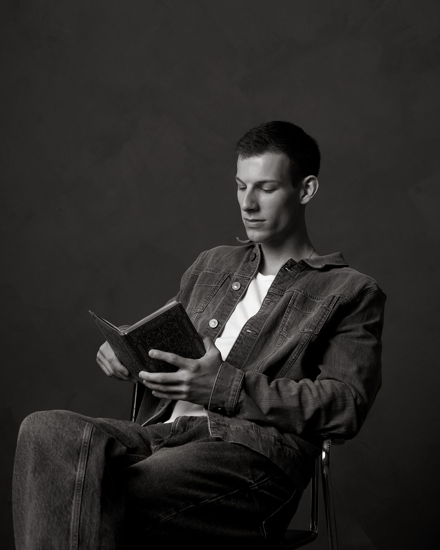 Young man in denim jacket sits reading a book, captured in classic black and white studio portrait photography.