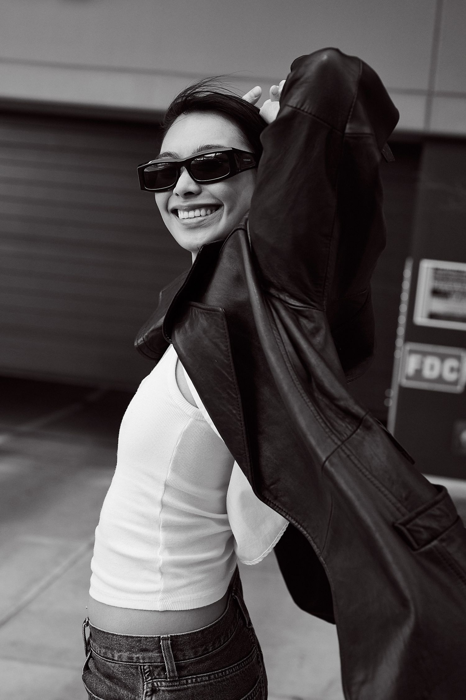 Smiling woman in sunglasses and white top holding black leather coat outdoors in black and white photo.