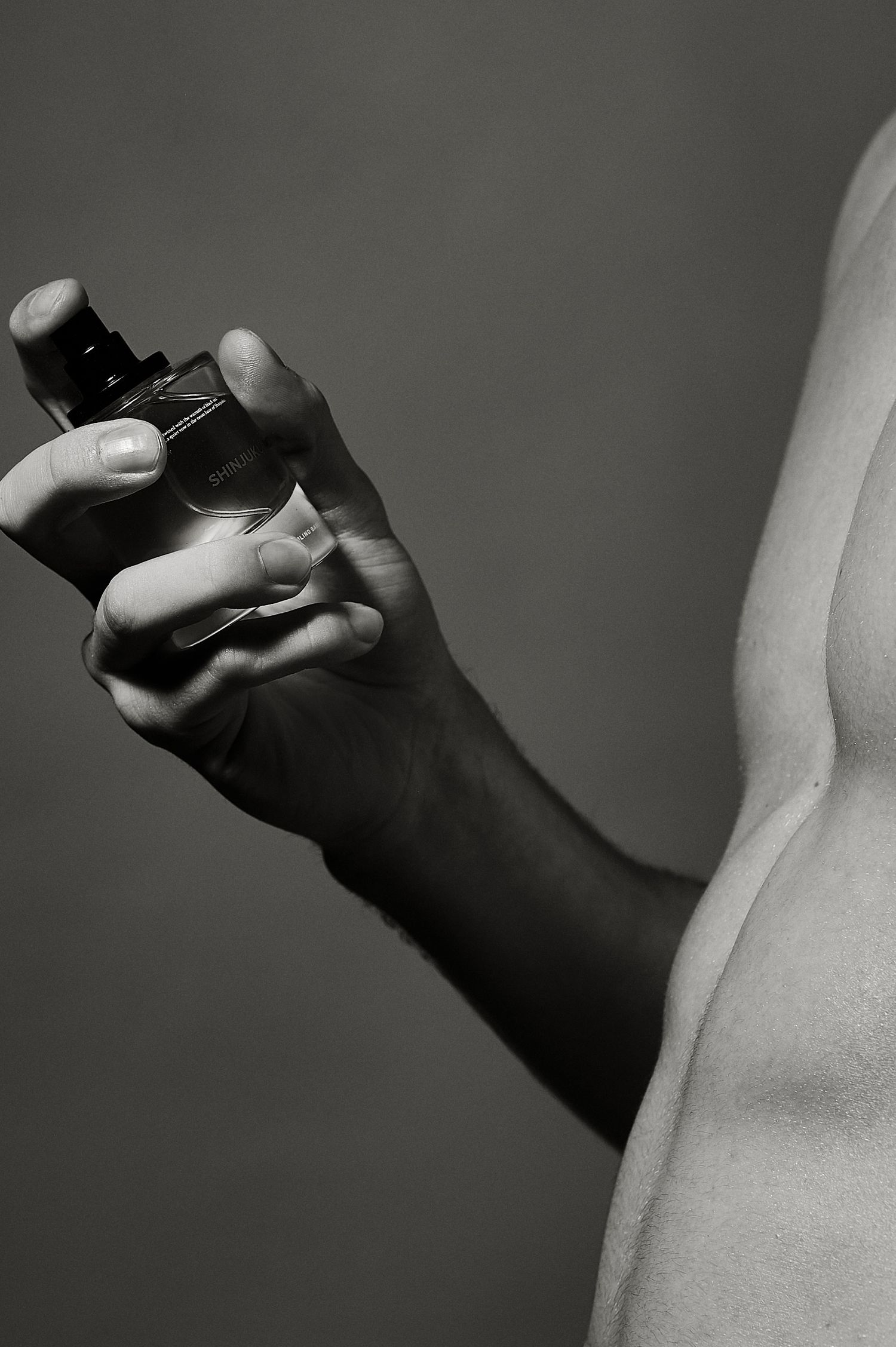 Close-up black and white photo of a hand holding a perfume bottle against a bare torso.
