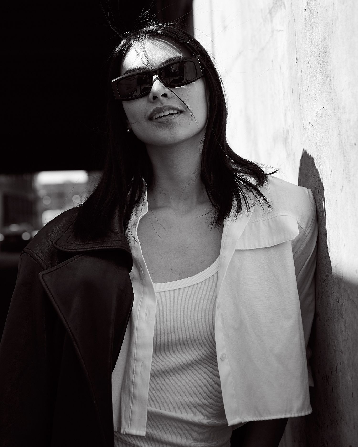 Black and white photo of dark-haired woman in sunglasses and leather jacket leaning against wall.