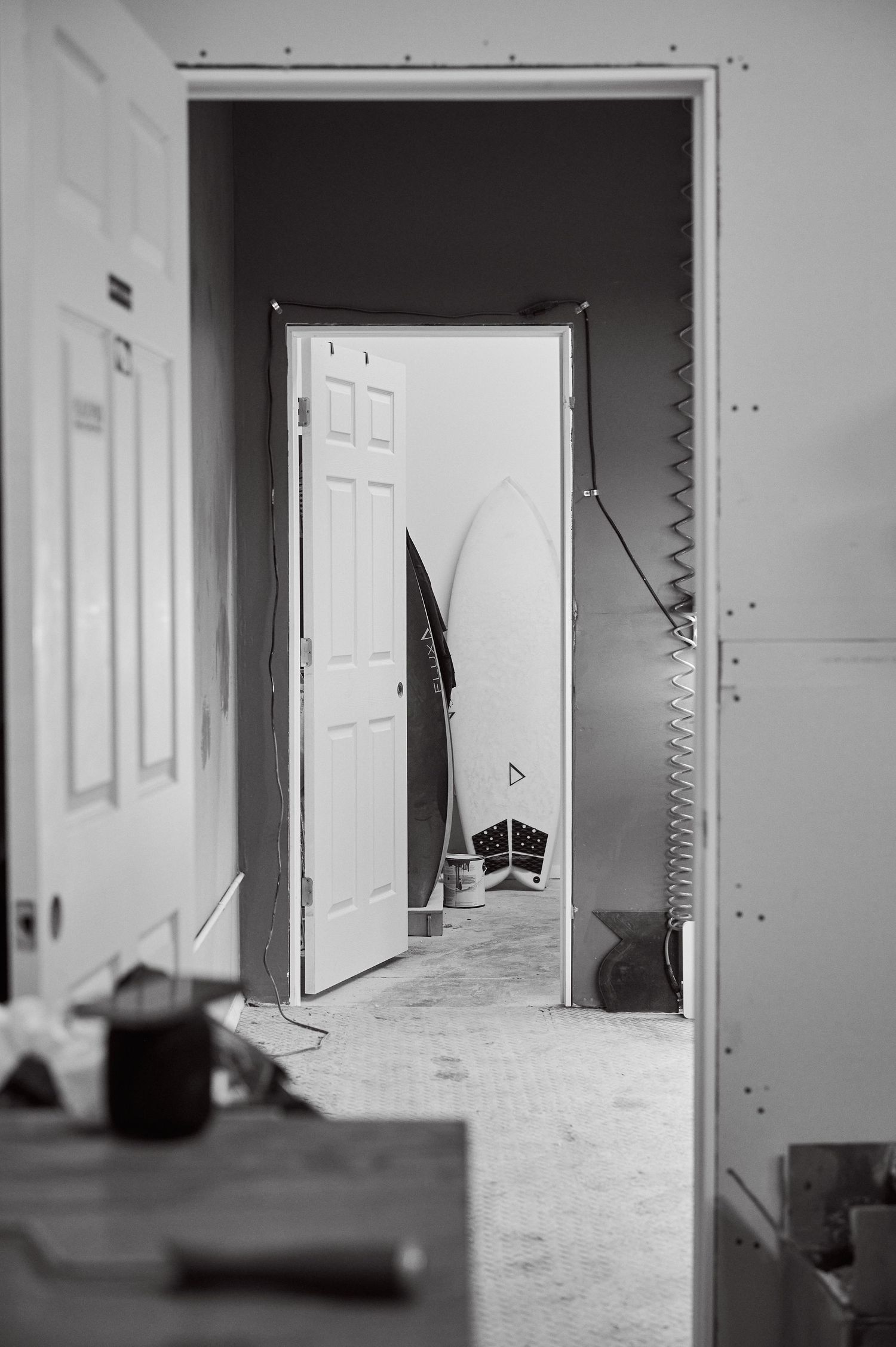 Black and white photo of surfboard visible through open doorway in workshop.