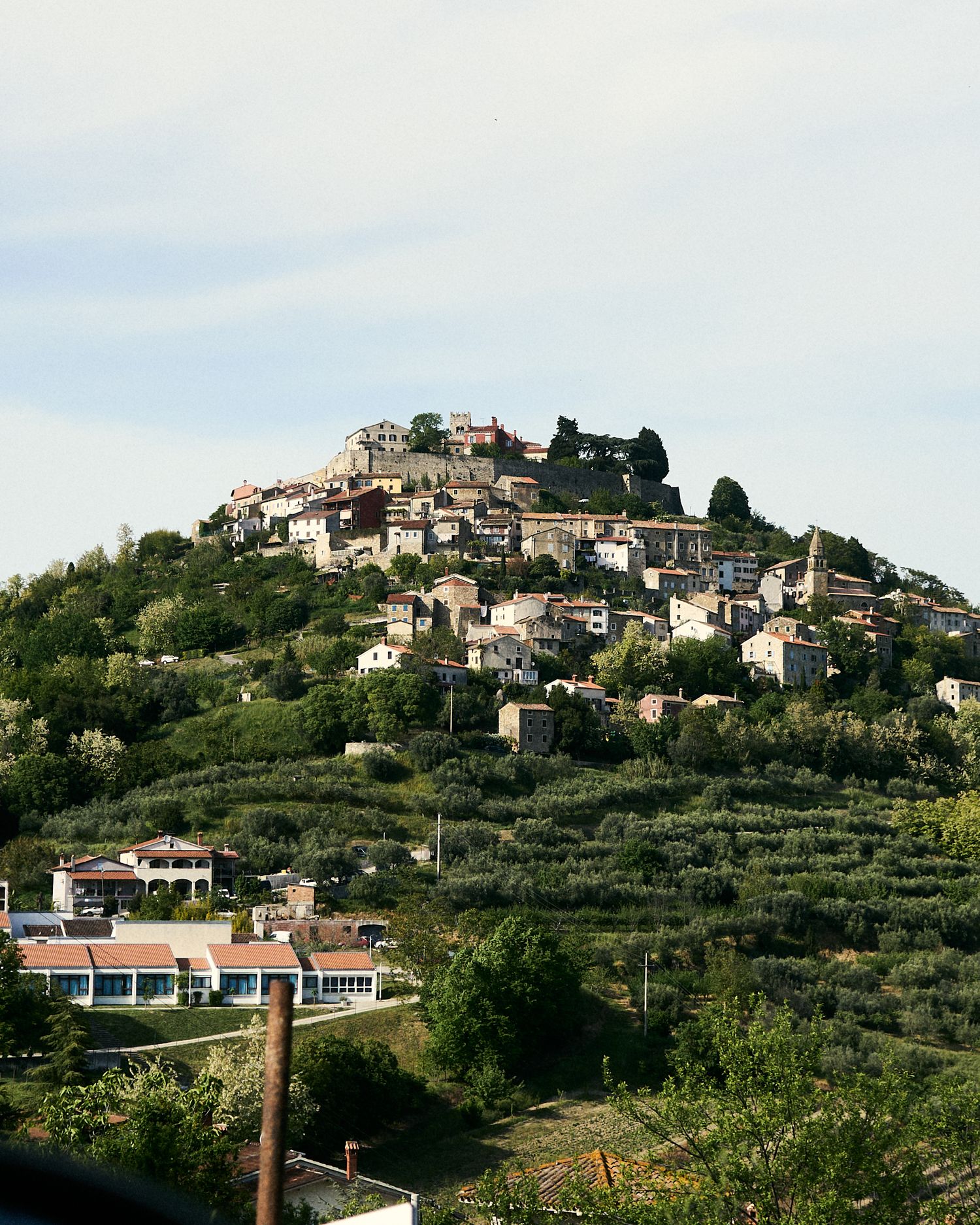 Hilltop medieval village with stone buildings surrounded by lush green trees and terraced hillsides under a cloudy sky.