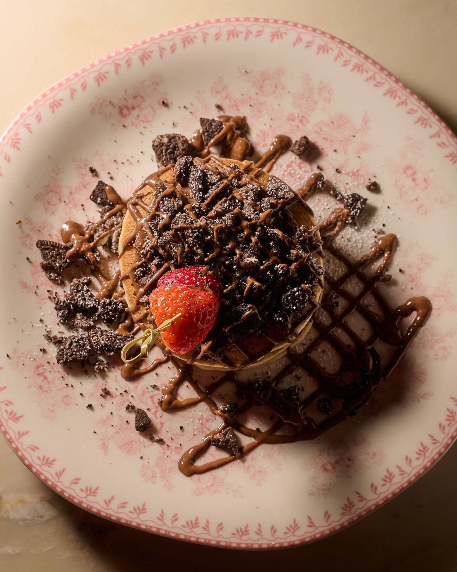 Chocolate waffle topped with shaved chocolate, caramel drizzle, and a fresh strawberry on a pink floral plate.