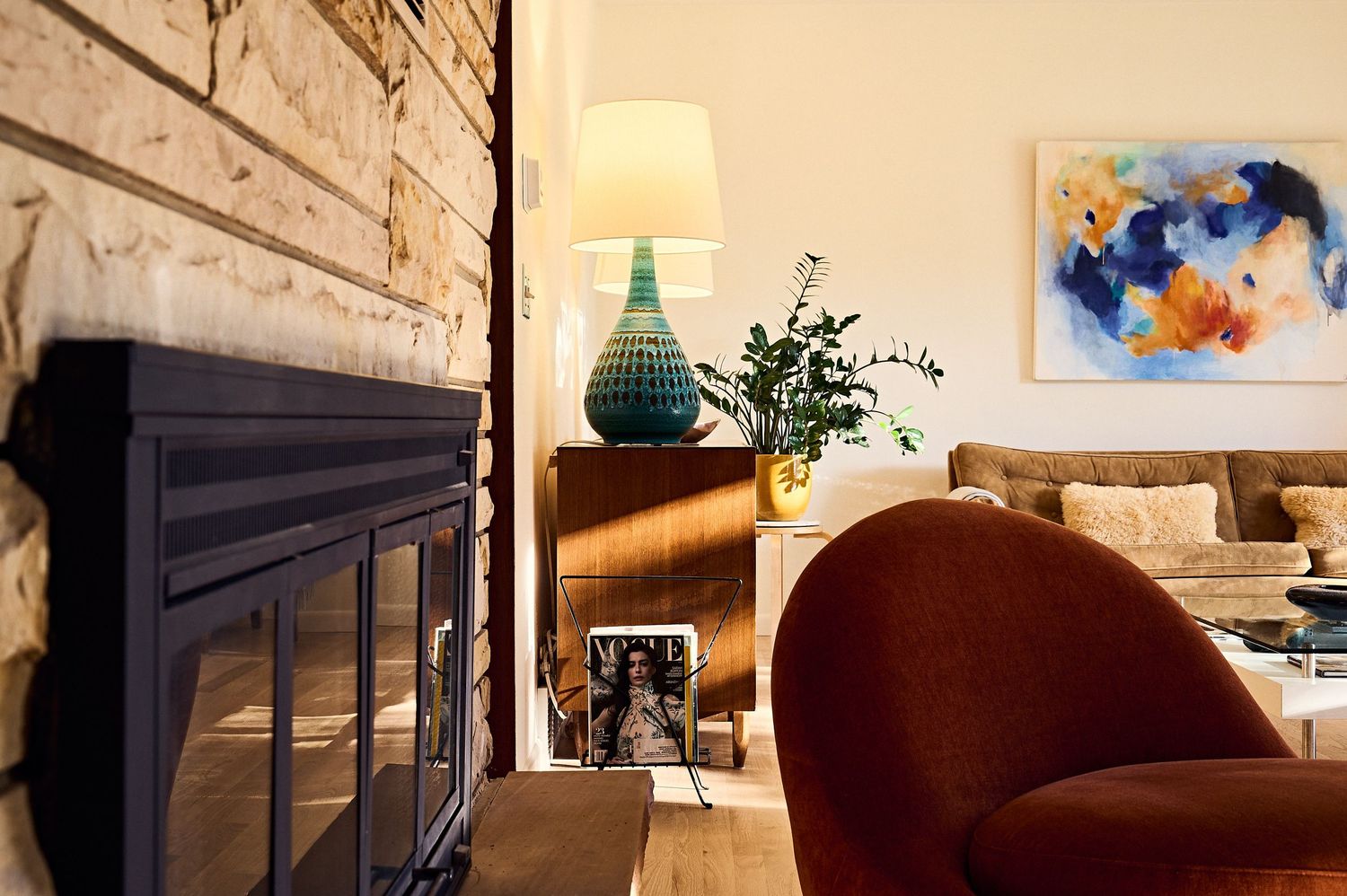 Cozy living room with brick fireplace, teal lamp, abstract wall art, and rust velvet chair in warm modern decor.
