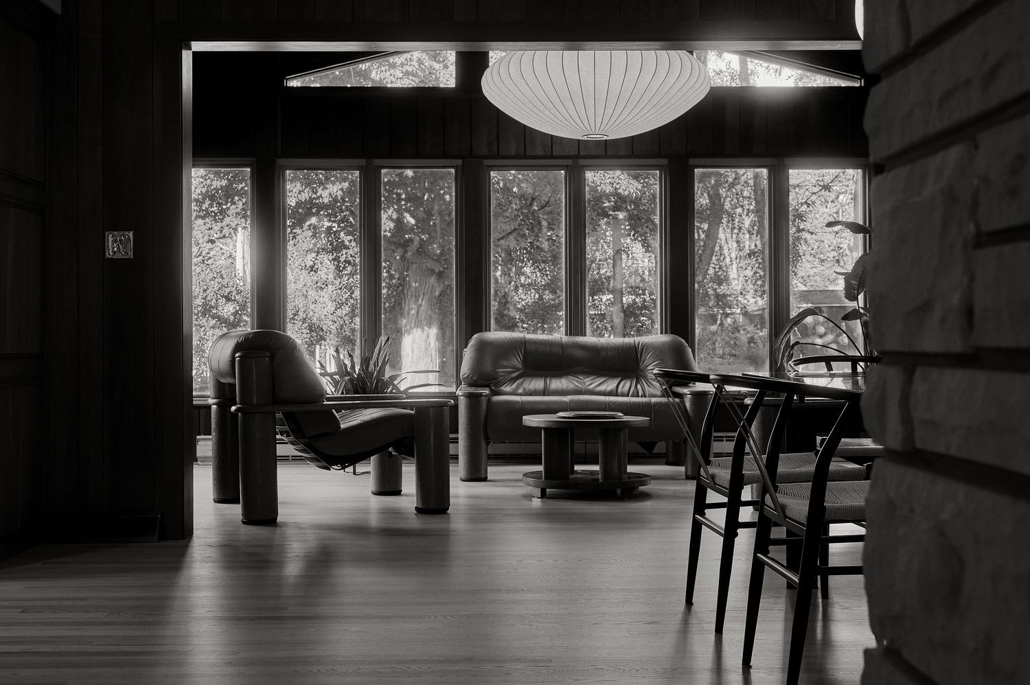 Black and white photo of a mid-century modern living room with leather chairs, a round pendant lamp, and large windows.