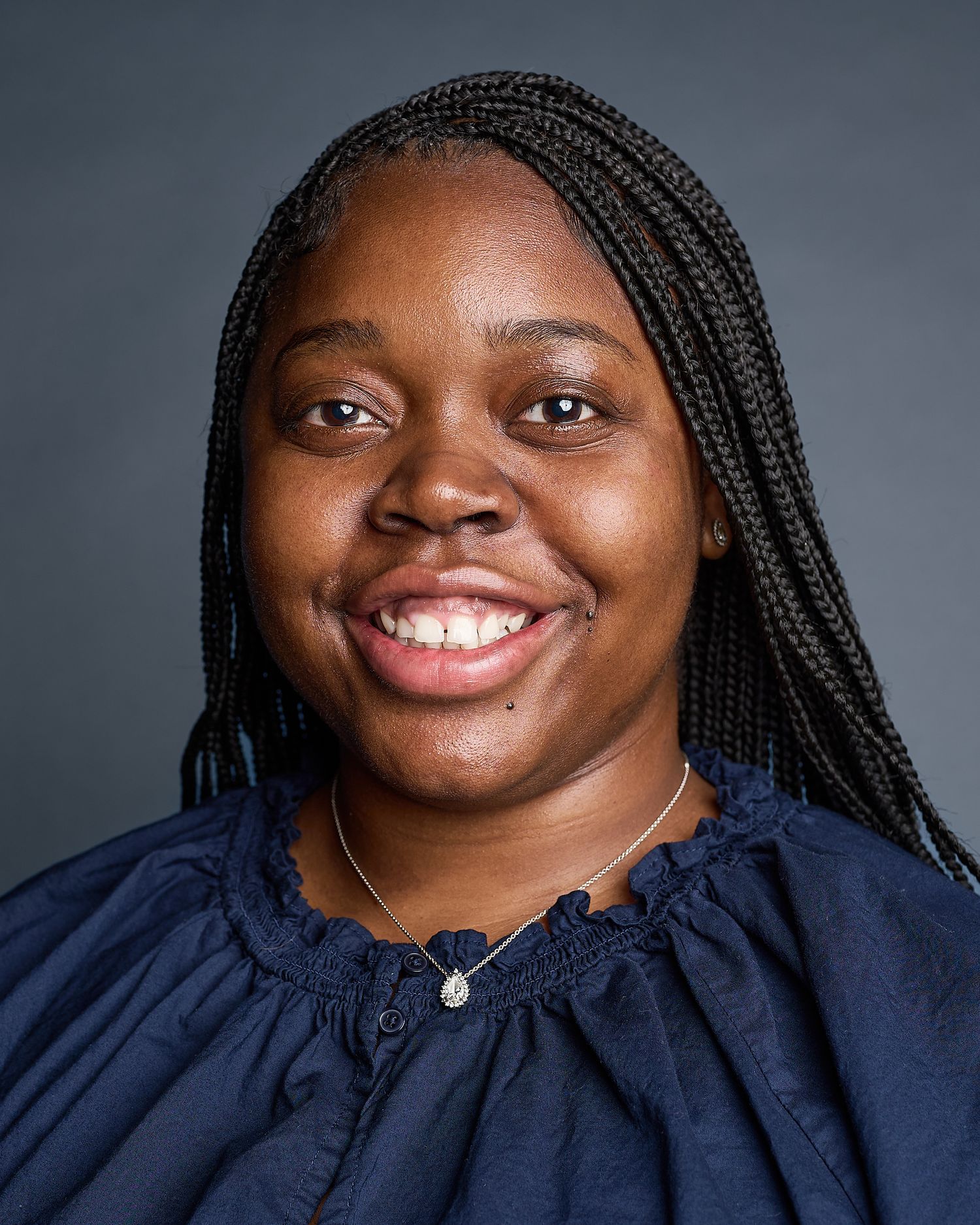 Smiling young woman with braided hair wearing a navy blue top and delicate necklace against a gray background.