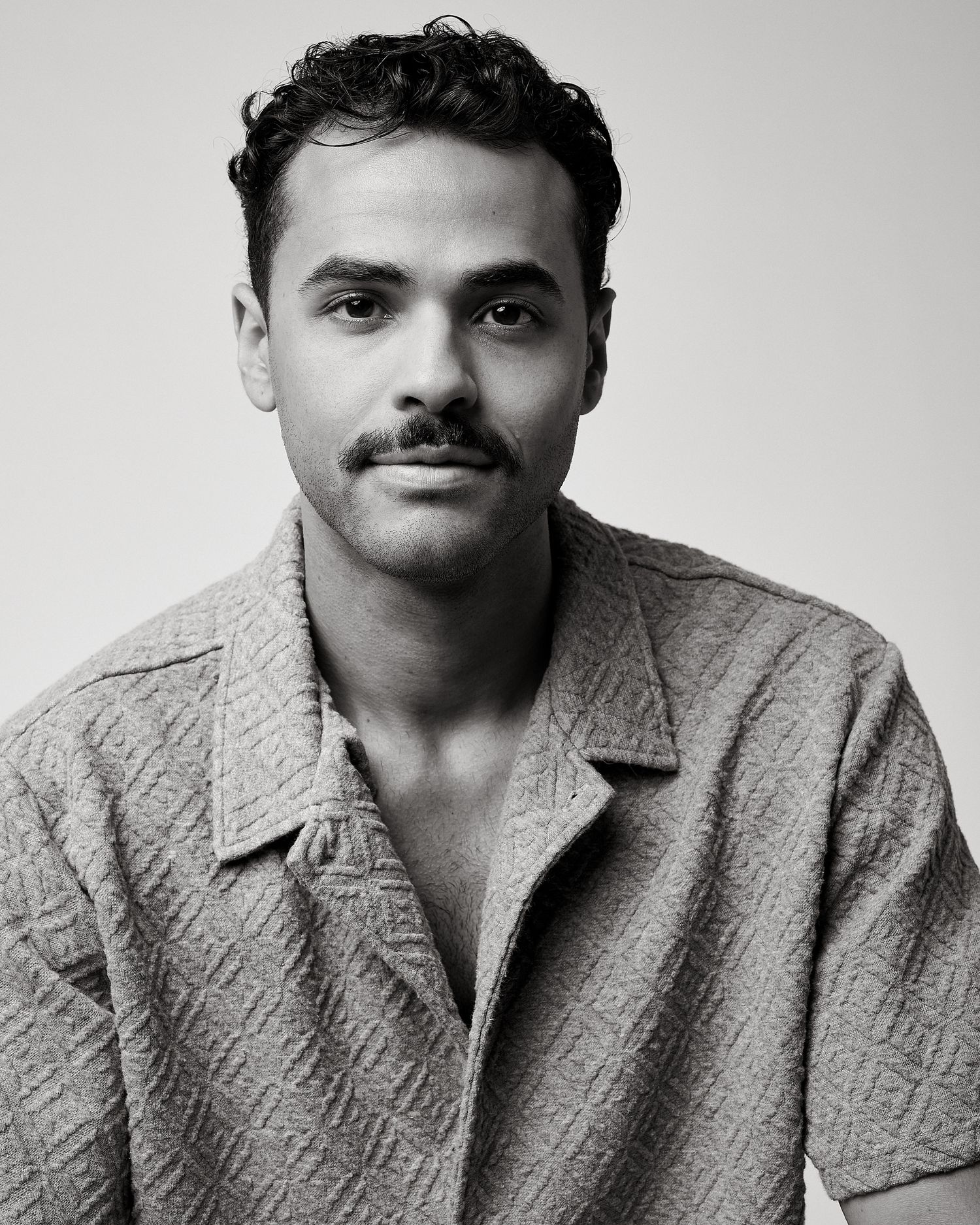 Black and white portrait of a young man with curly hair and mustache wearing a textured button-up shirt.
