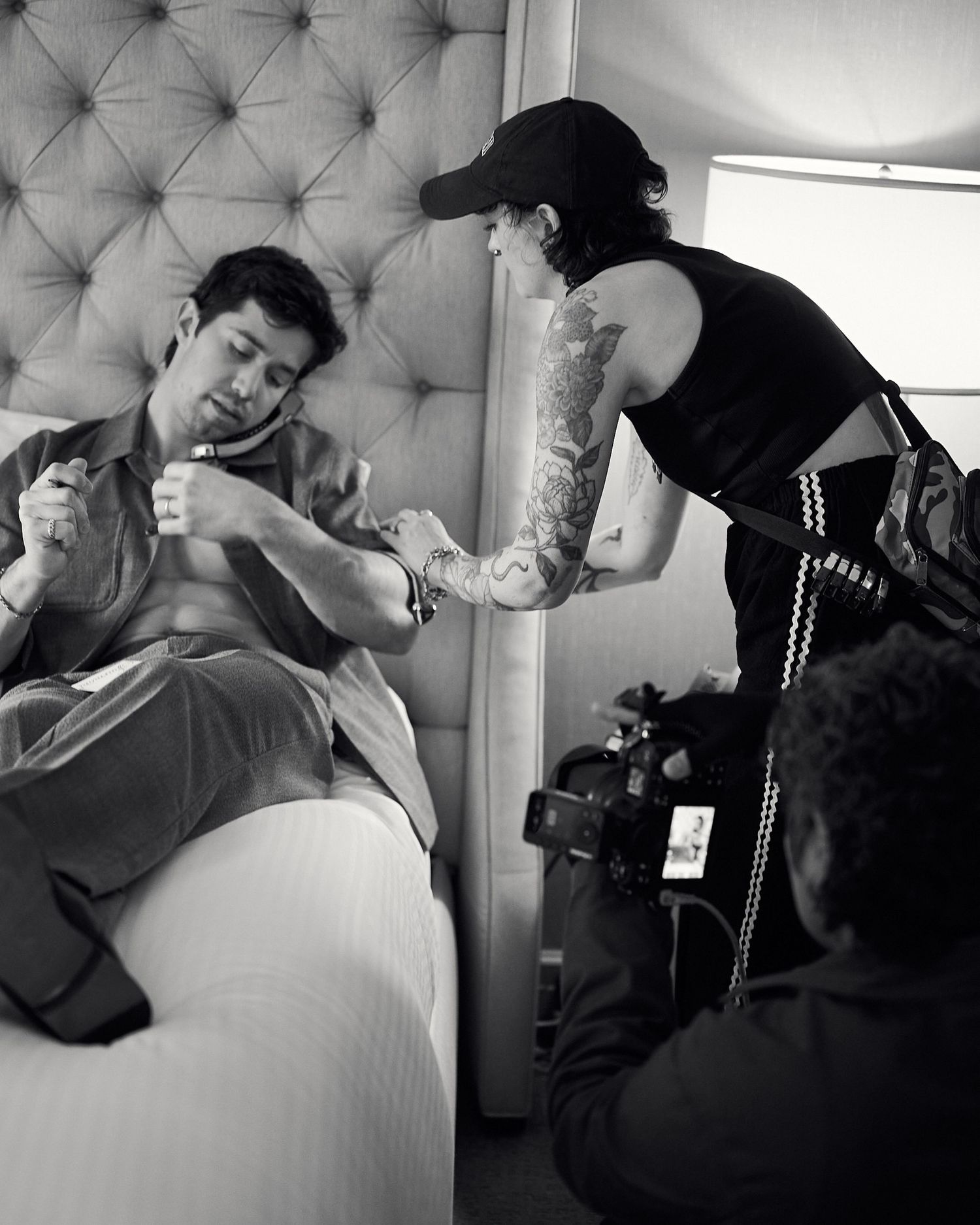Black and white behind-the-scenes photo of a shirtless man on a bed being styled by a tattooed crew member.