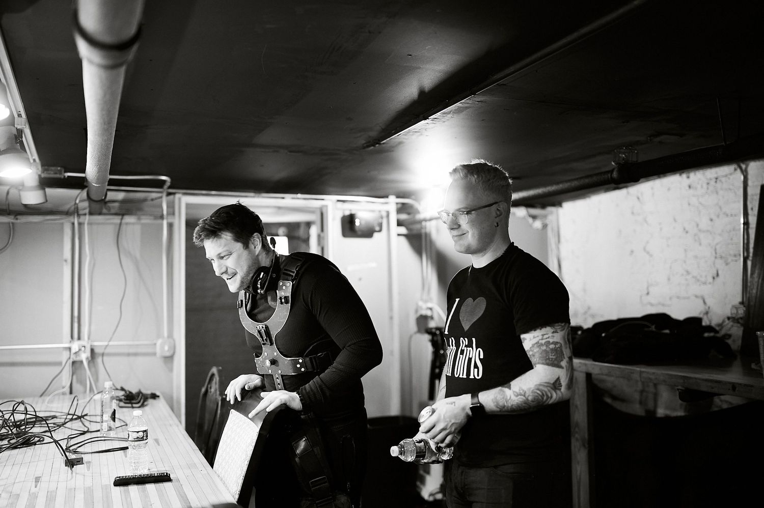 Two men working in a dimly lit backstage area with audio equipment in a black and white photo.