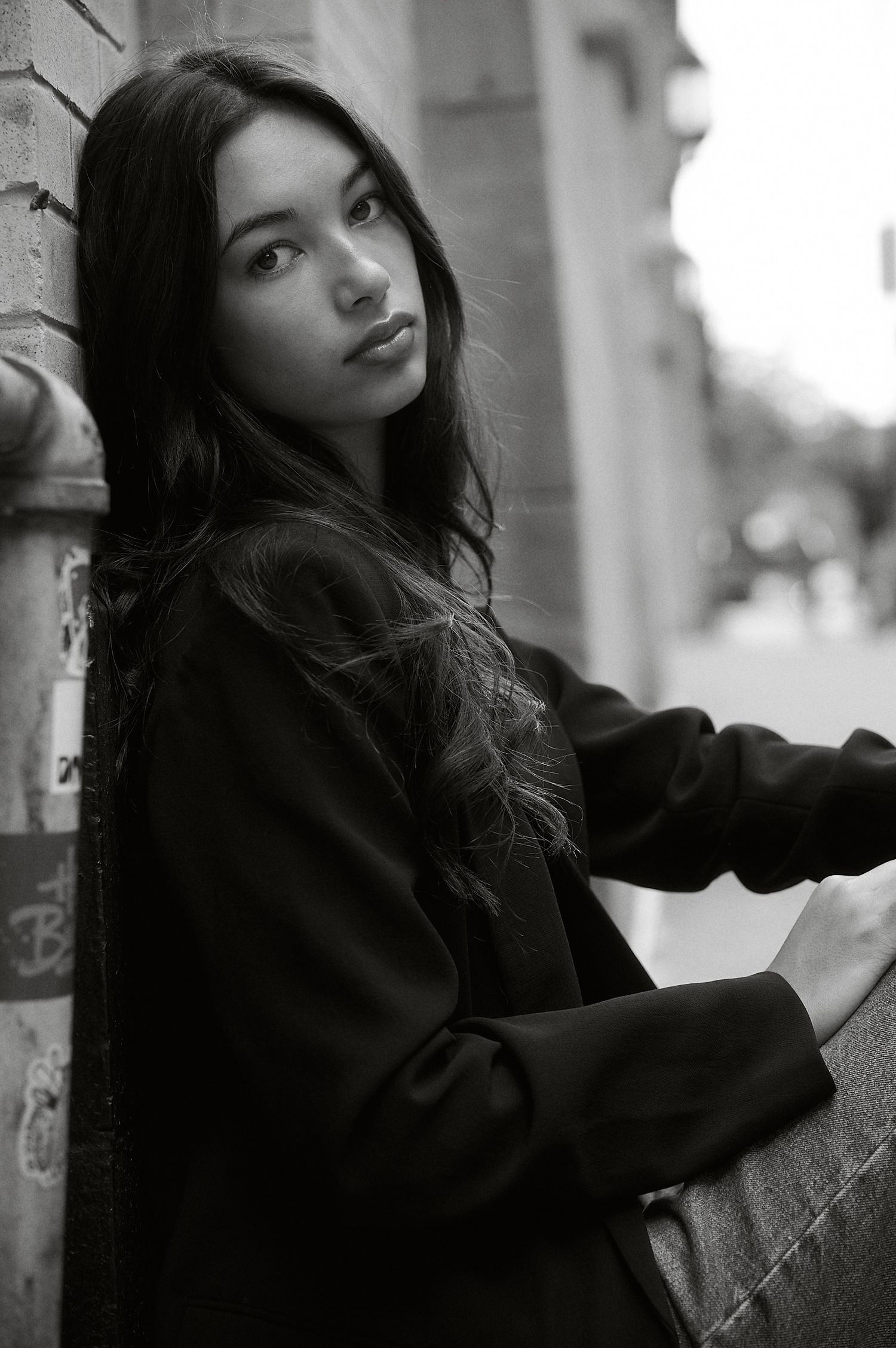 Black and white portrait of a young woman with long dark hair leaning against a brick wall outdoors.