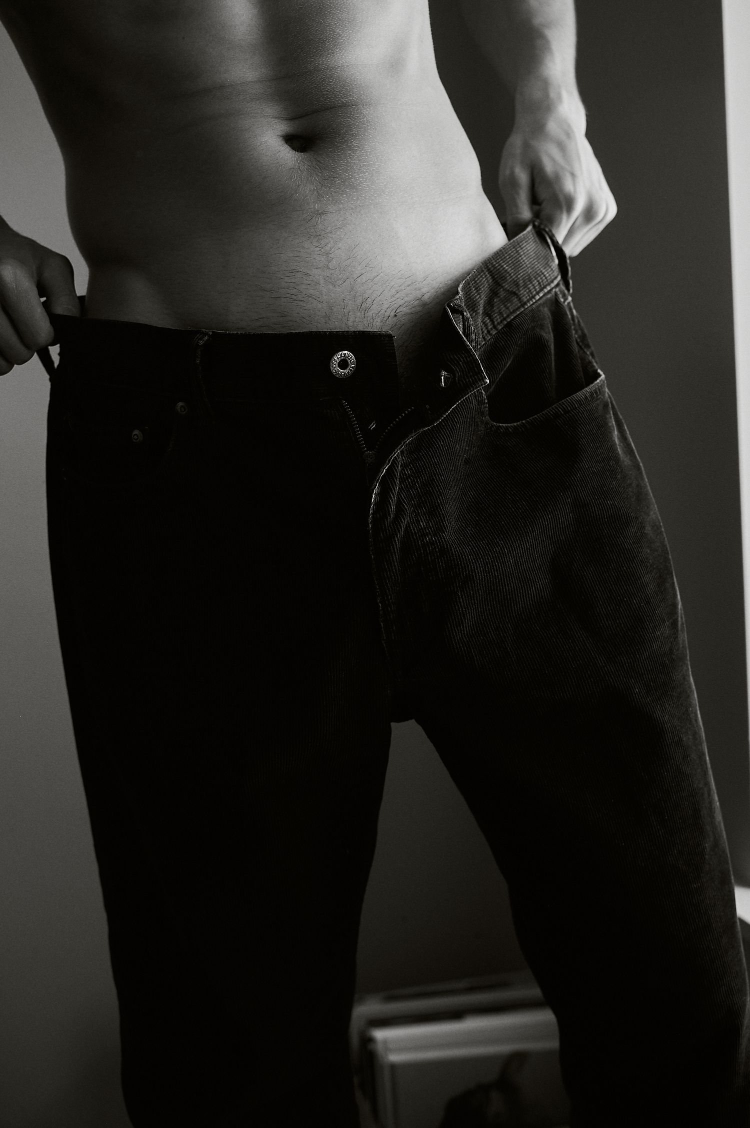 Black and white photo of a shirtless muscular man in dark jeans, showing toned abs and athletic physique.