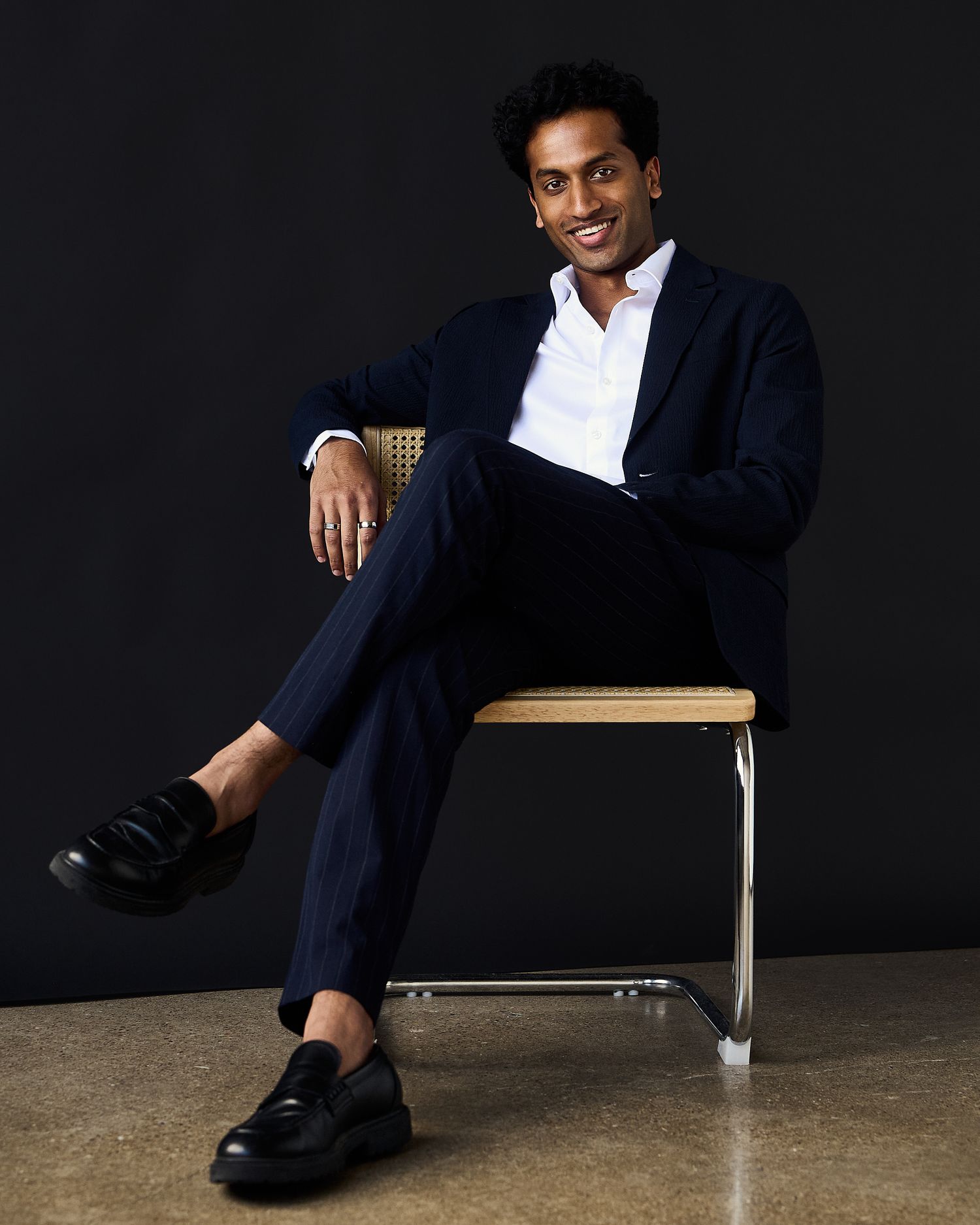 A stylish man in a navy suit and white shirt sits cross-legged on a chair, smiling against a dark background.