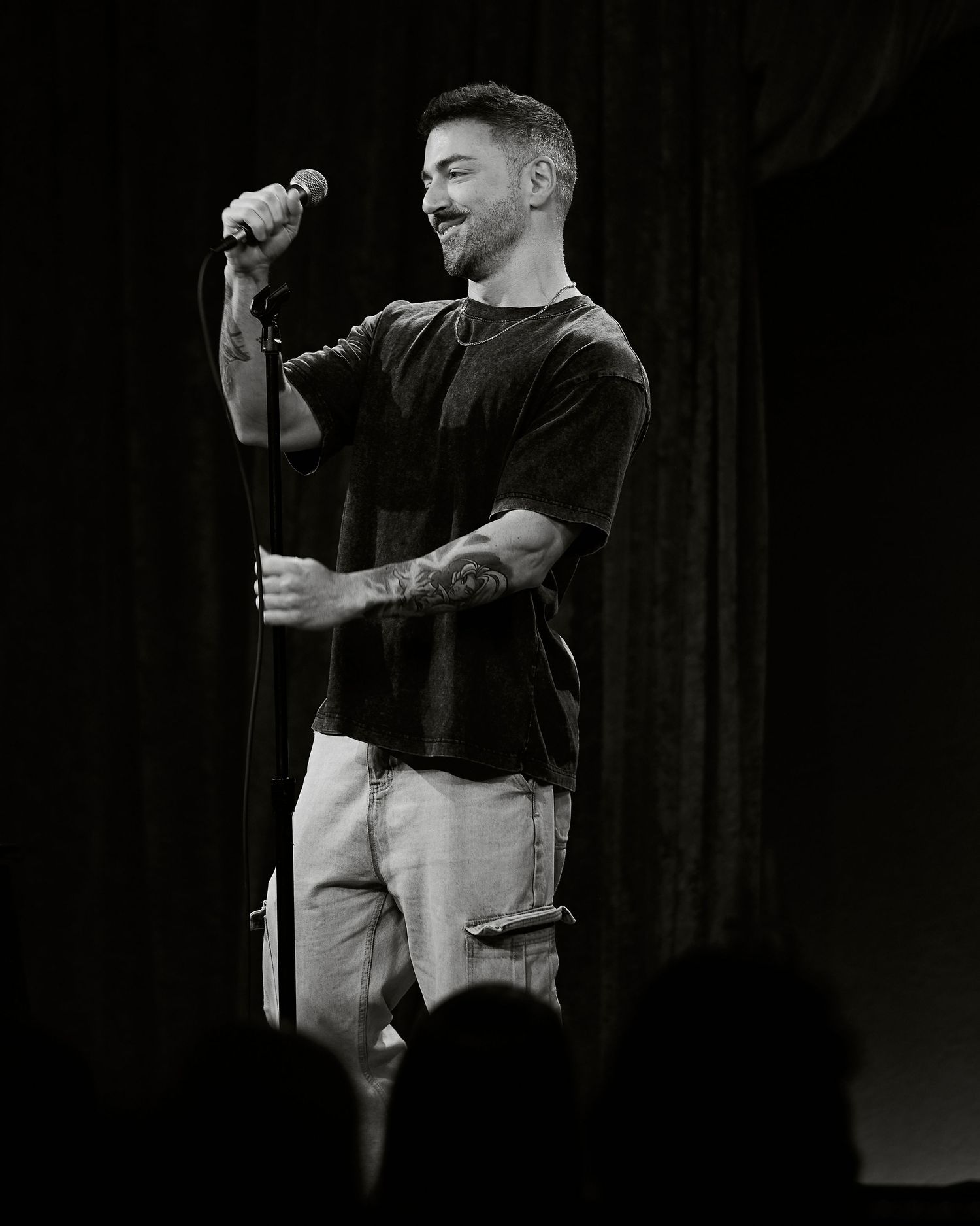 Black and white photo of a tattooed man smiling while performing on stage with a microphone.