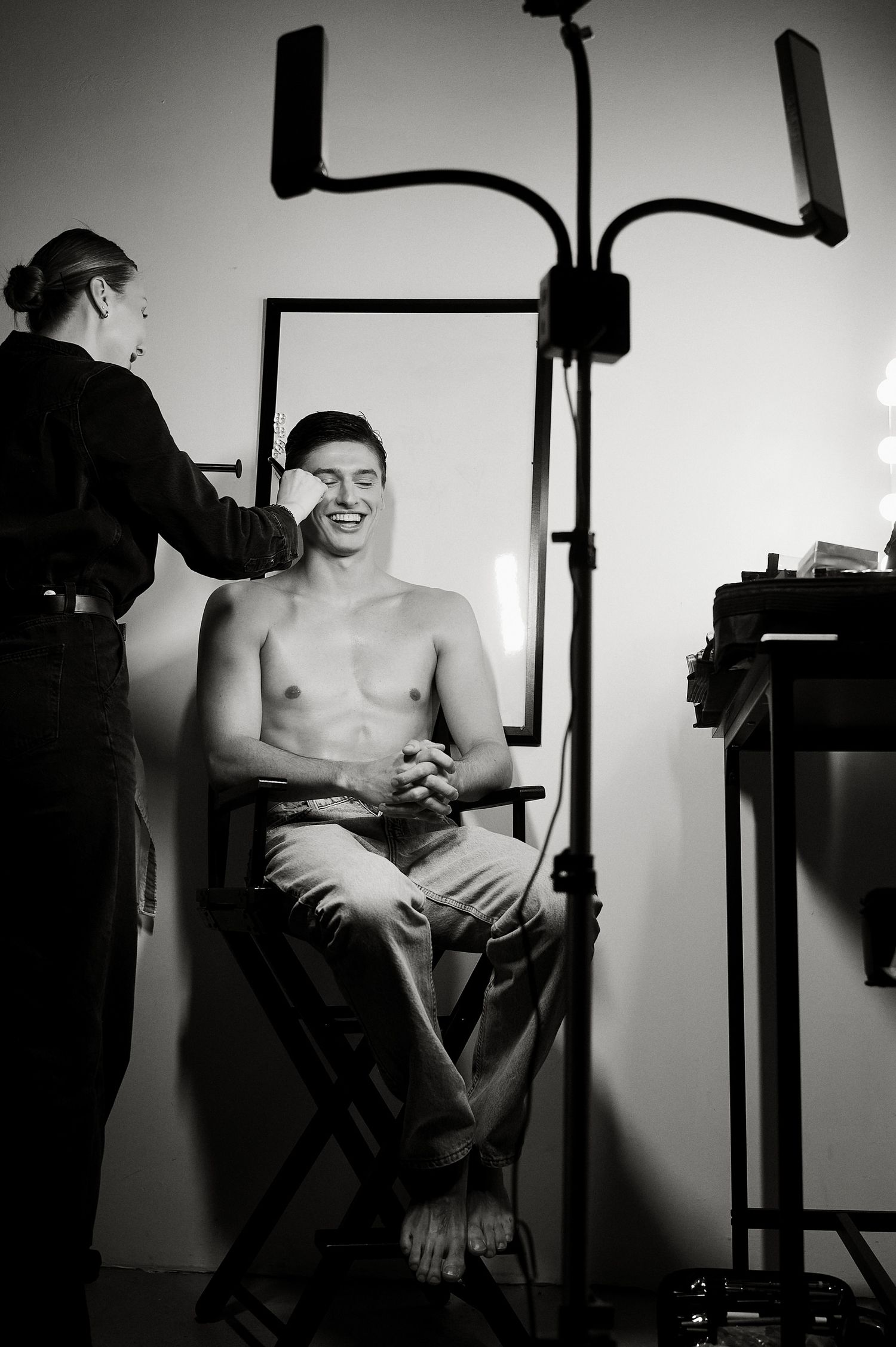 Black and white behind-the-scenes photo of a makeup artist applying makeup to a smiling shirtless male model.