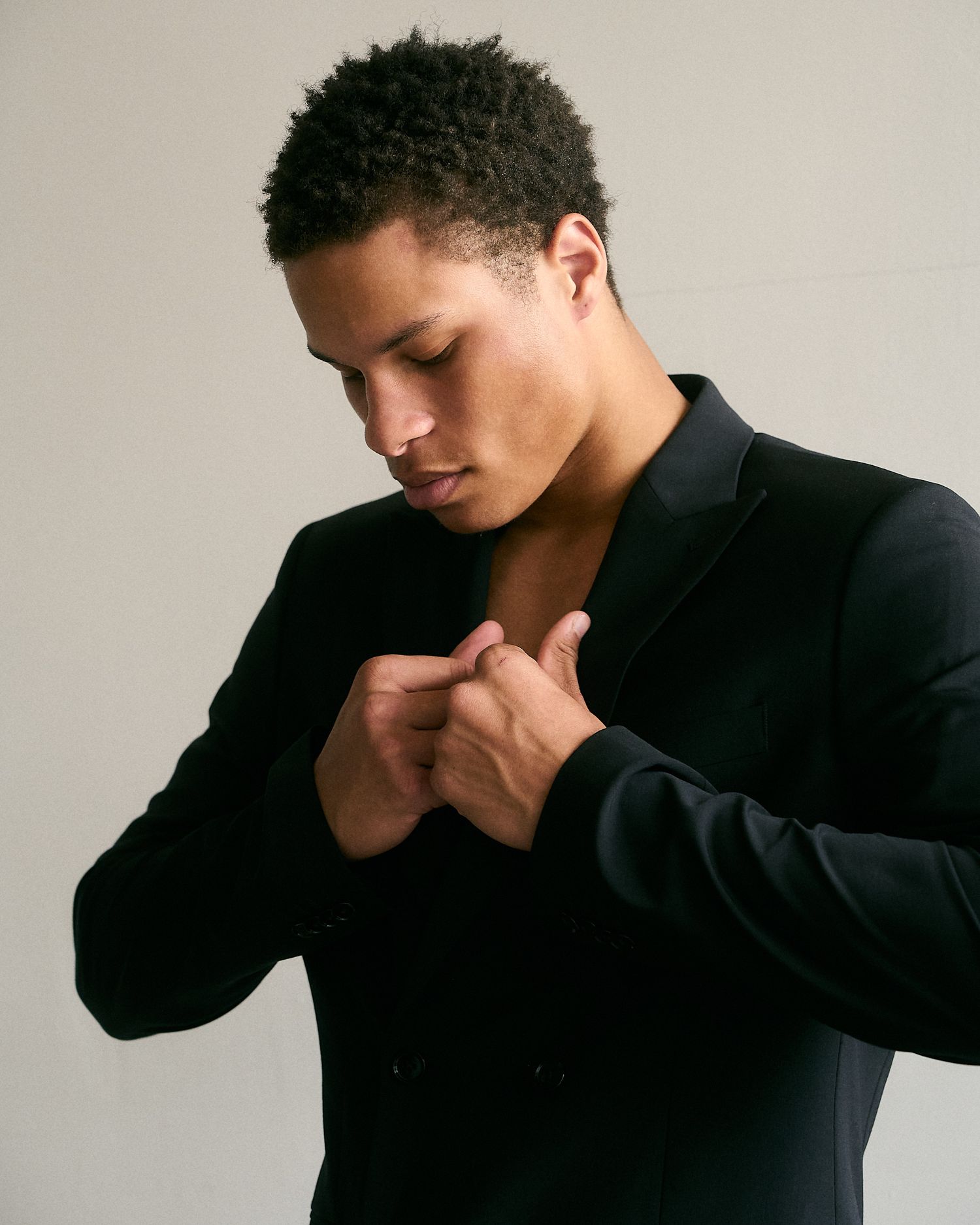 Young man in black blazer looking down with hands clasped against chest on neutral background.