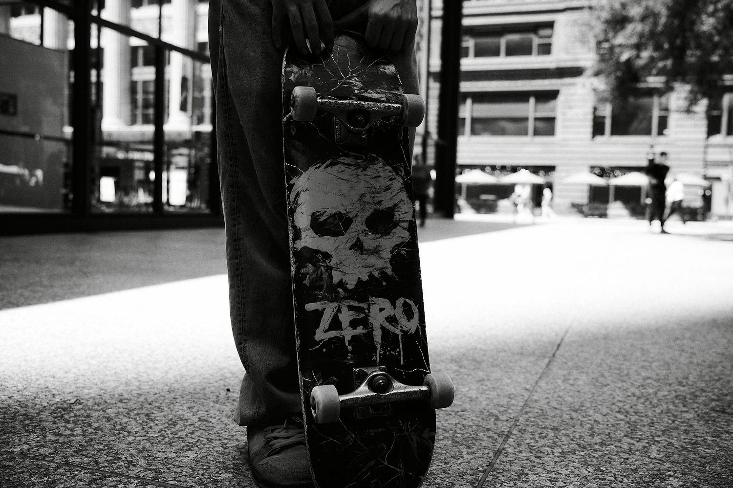 Black and white photo of a person holding a Zero skateboard with skull graphic on a city sidewalk.