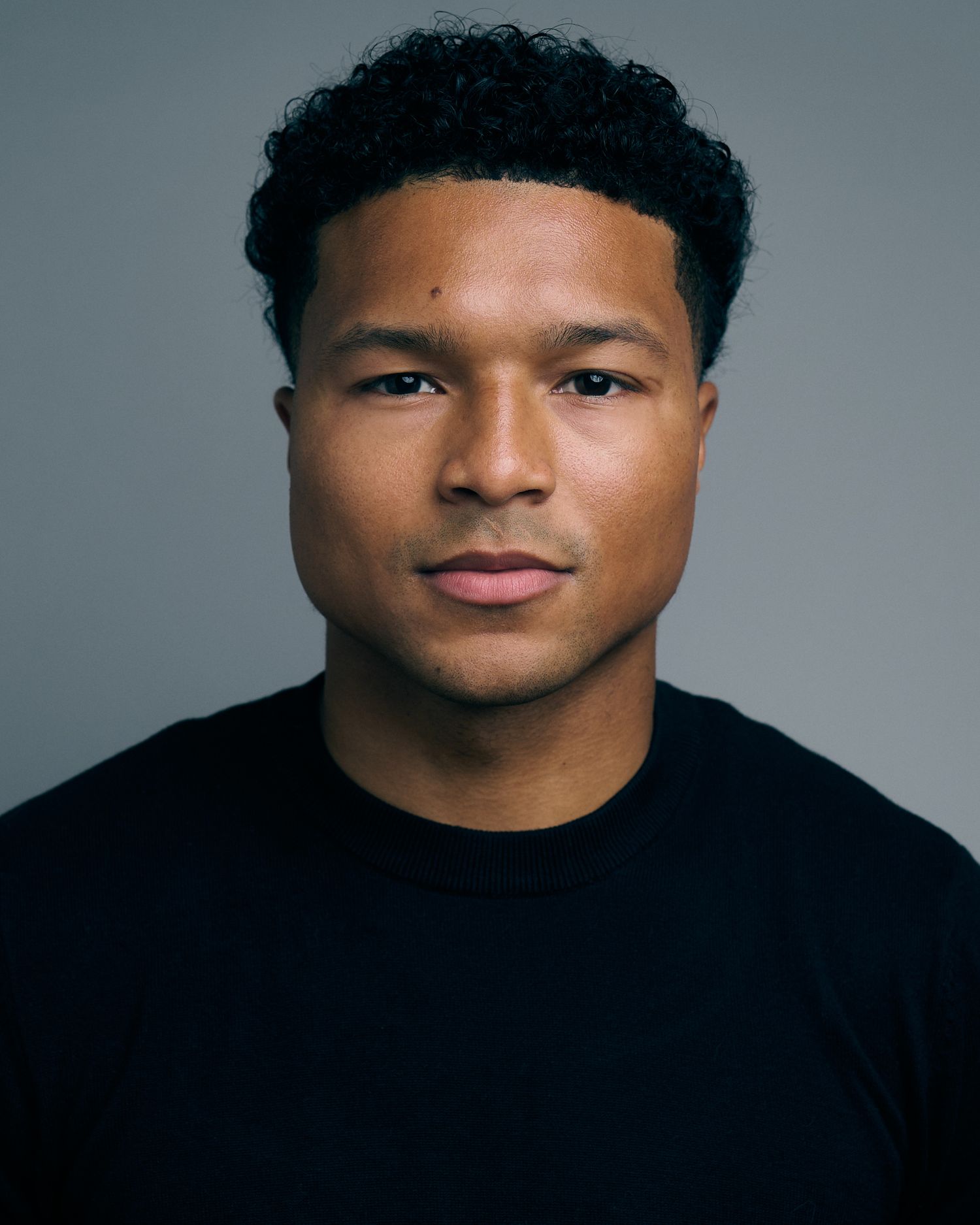 Young Black man with curly hair wearing a black crew neck shirt poses for a professional headshot photo.