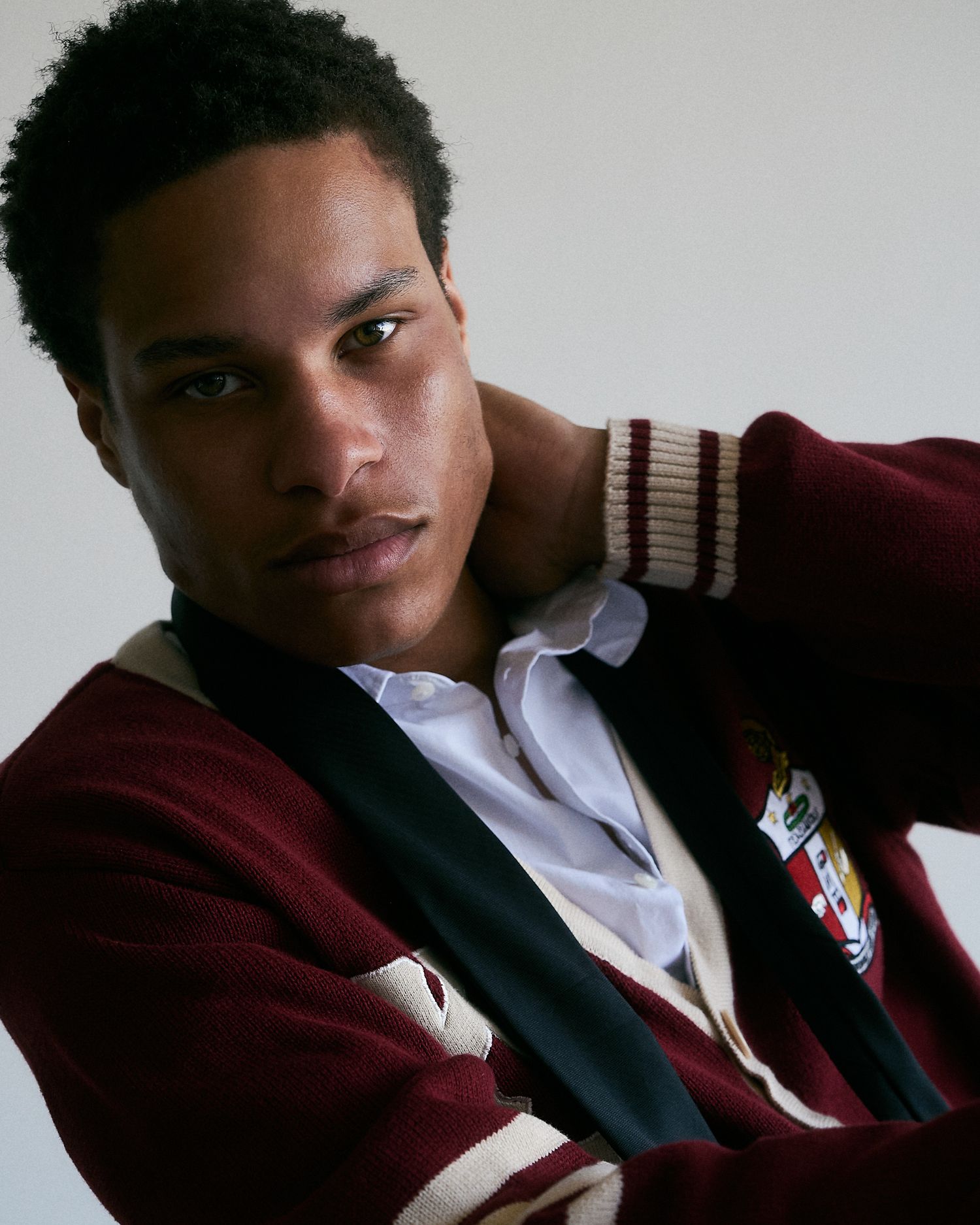 Young man in burgundy varsity cardigan with crest patch poses confidently against white background.