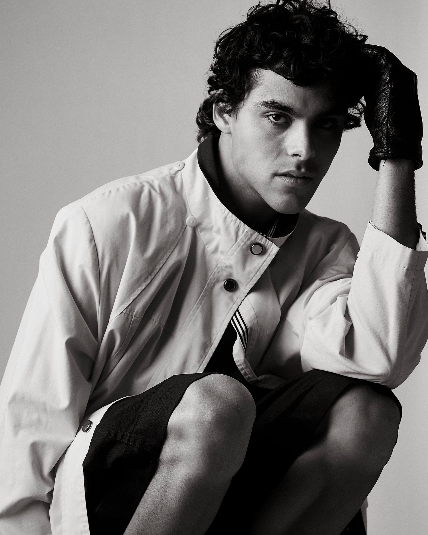 Black and white portrait of a young male model with curly hair in a white shirt seated casually.