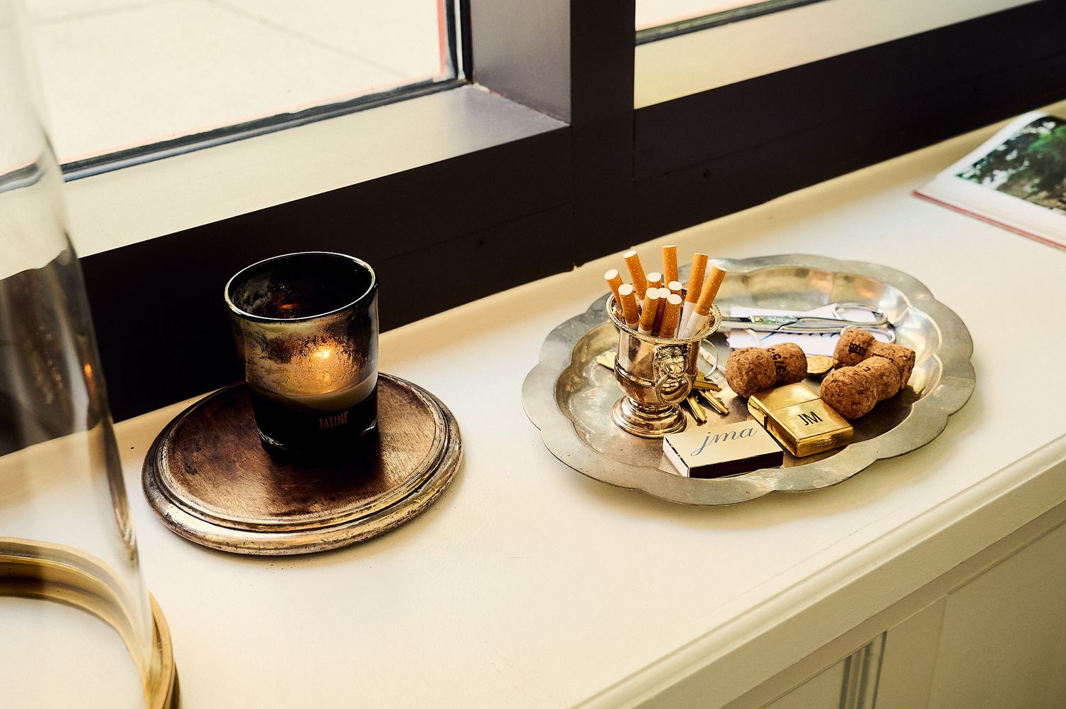 Silver tray with croissants and snacks beside a dark glass on a wooden coaster near a window.