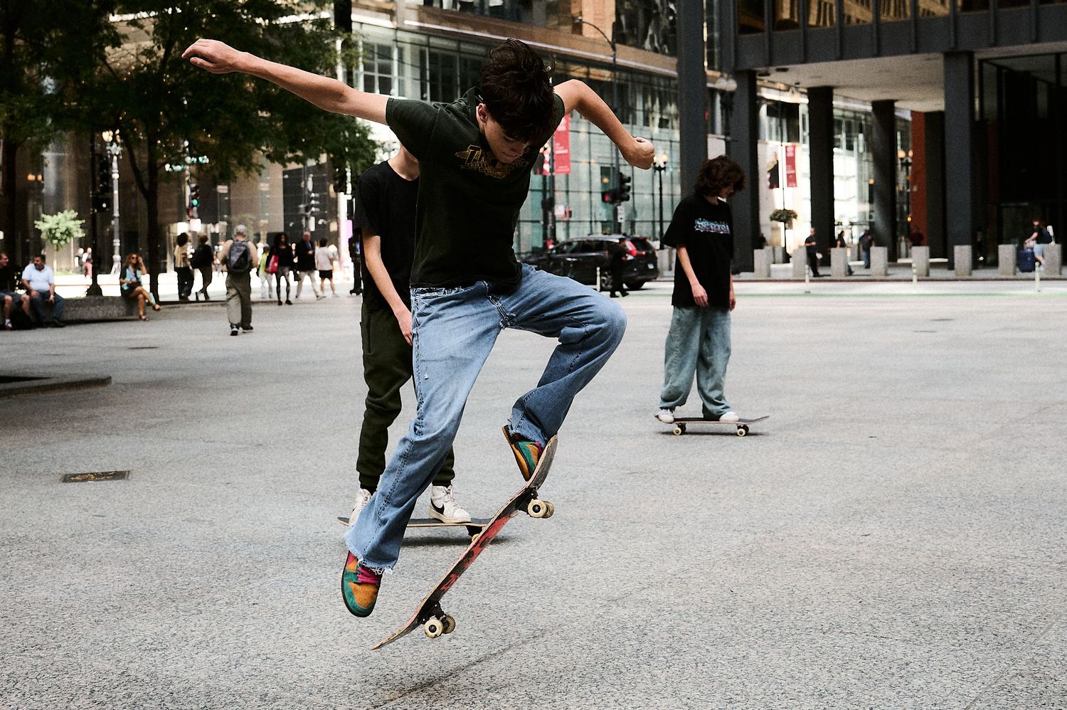 A skateboarder performs a kickflip trick in an urban plaza surrounded by city buildings and onlookers.