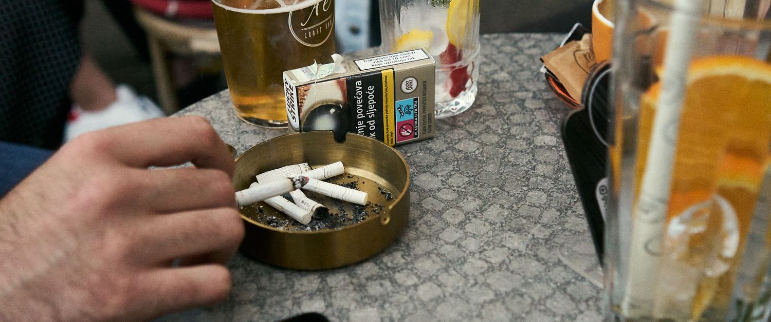 Hand placing cigarettes in ashtray on table with beer glasses and cigarette pack in bar setting.