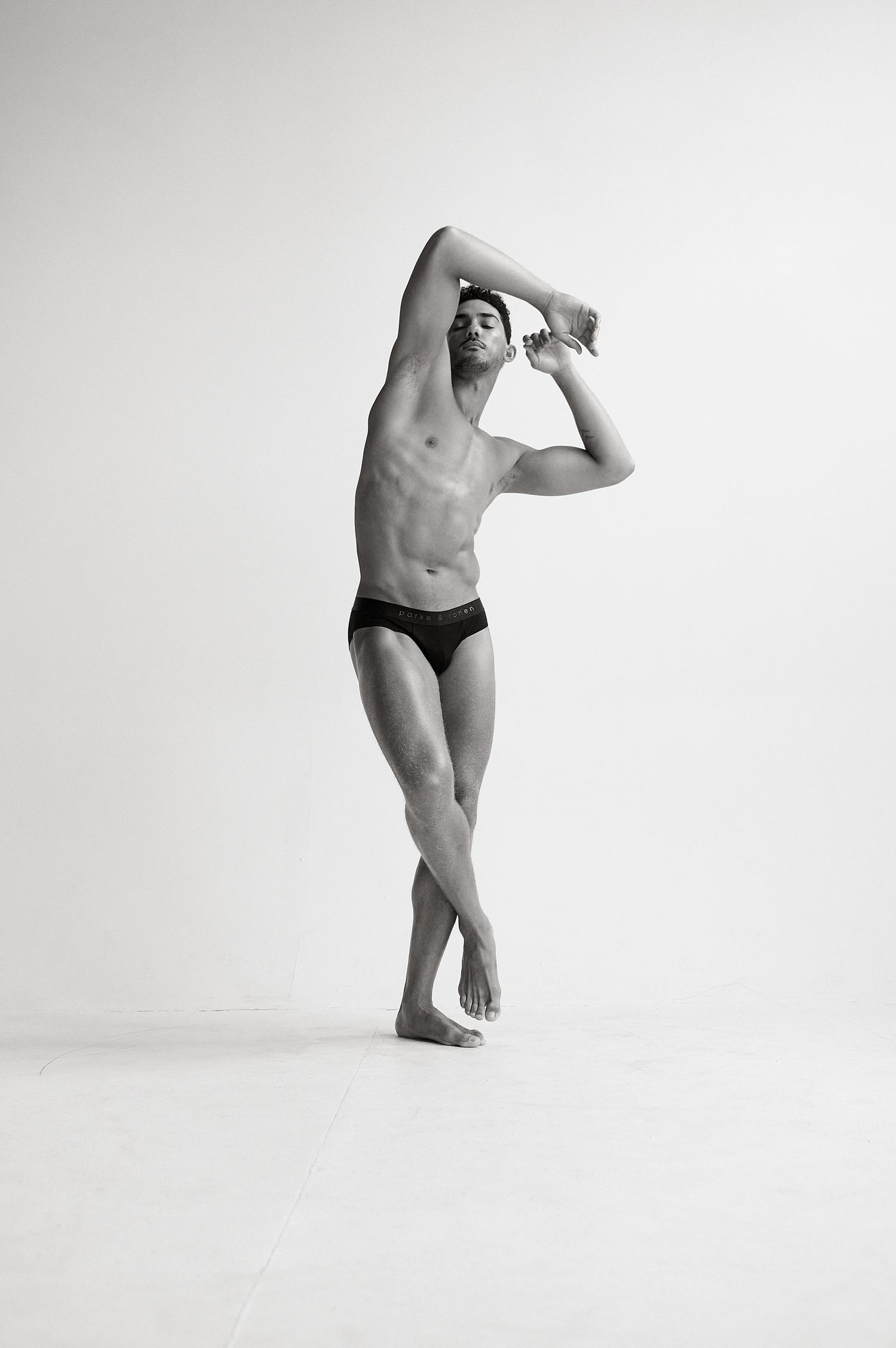 Shirtless muscular male dancer stretching arms overhead in black briefs in black and white studio shot.