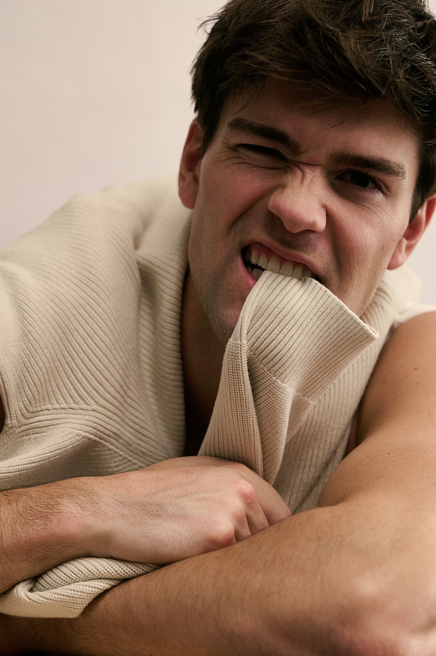 Young man in beige ribbed sweater biting his sleeve with arms crossed, making an intense facial expression.