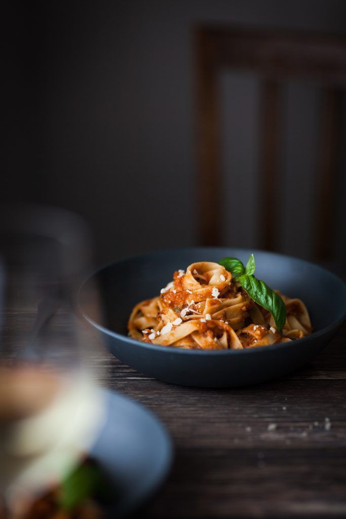A dark bowl of fettuccine pasta in tomato sauce, garnished with fresh basil and Parmesan, served on a rustic wooden table.