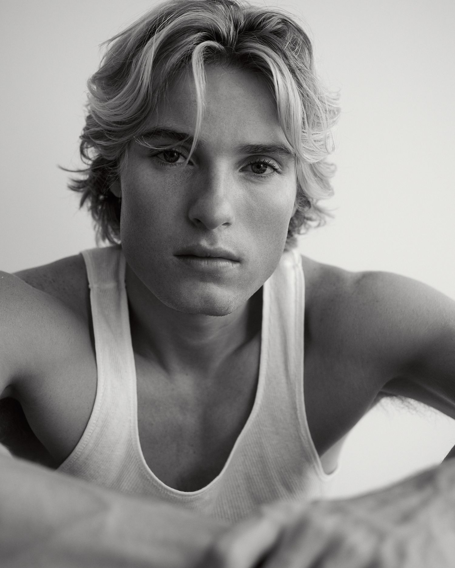 Black and white close-up of a young blonde male model in a white tank top leaning forward intensely.