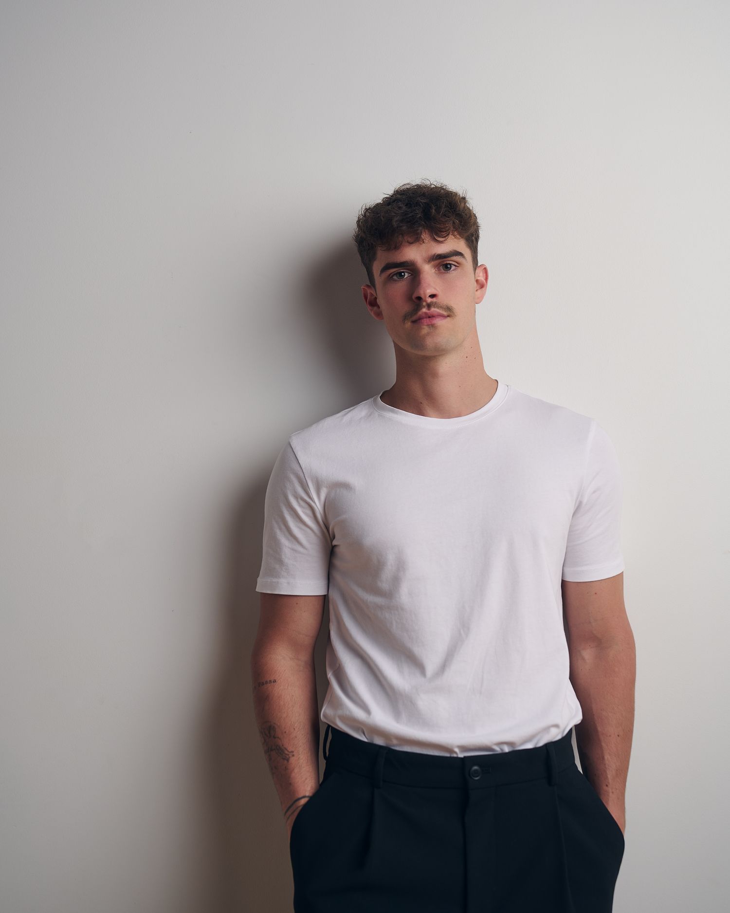 Young man with curly hair wearing a white t-shirt and dark trousers standing against a light wall.