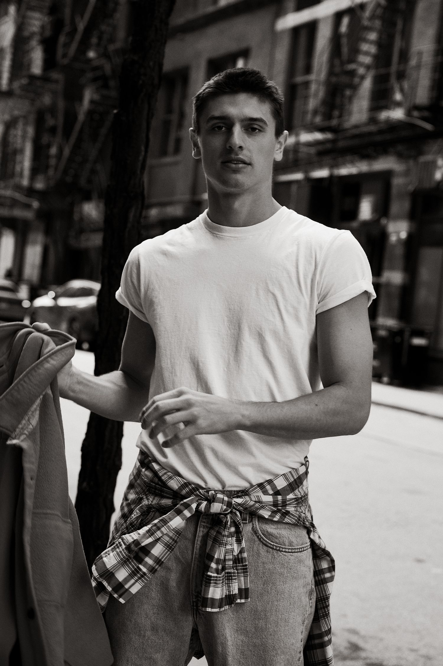 Young man in white tee and plaid shirt tied at waist poses on urban street in black and white photo.