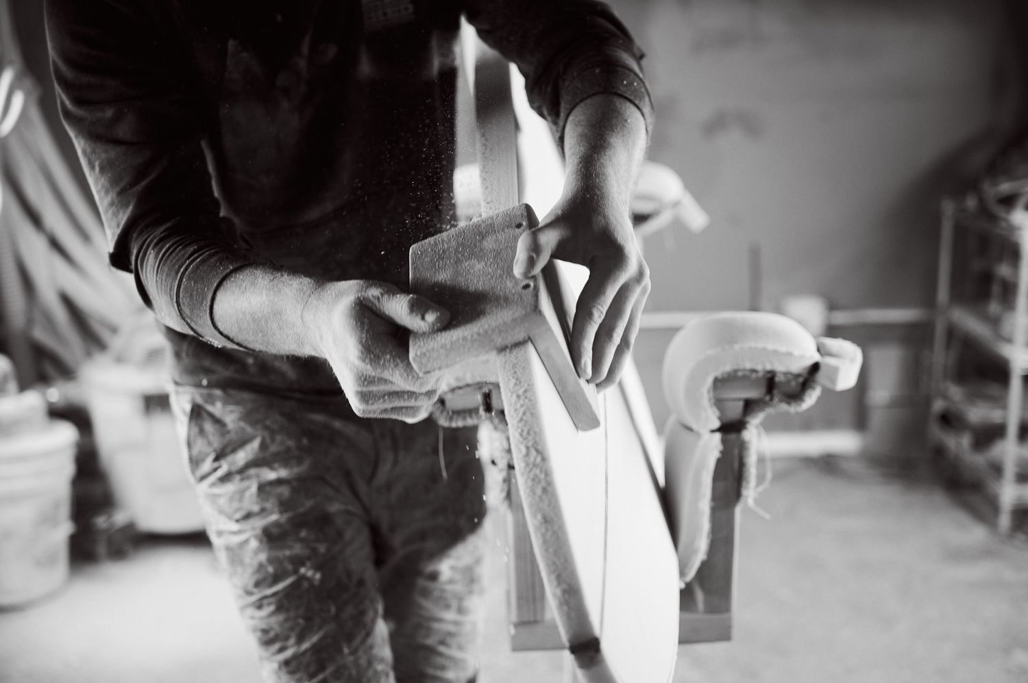 A craftsman uses a hand plane to shape a surfboard blank in a workshop.