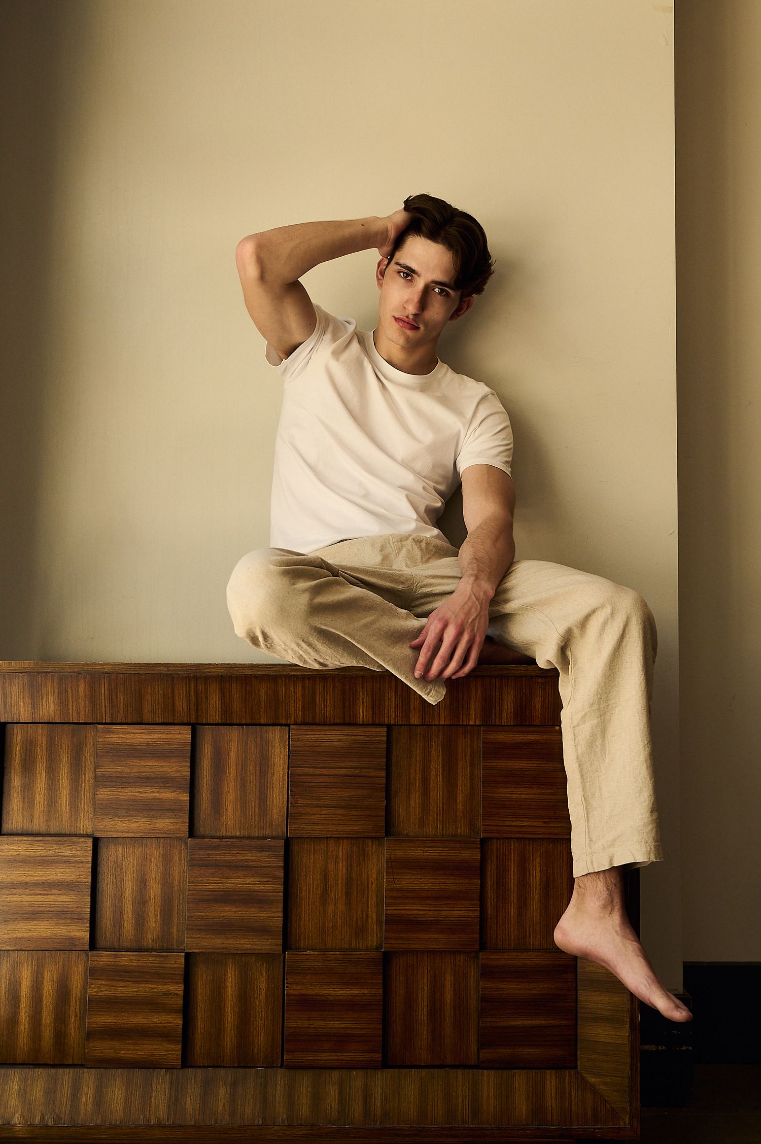 Young man in white tee and beige pants sitting barefoot on wooden cabinet against neutral wall.