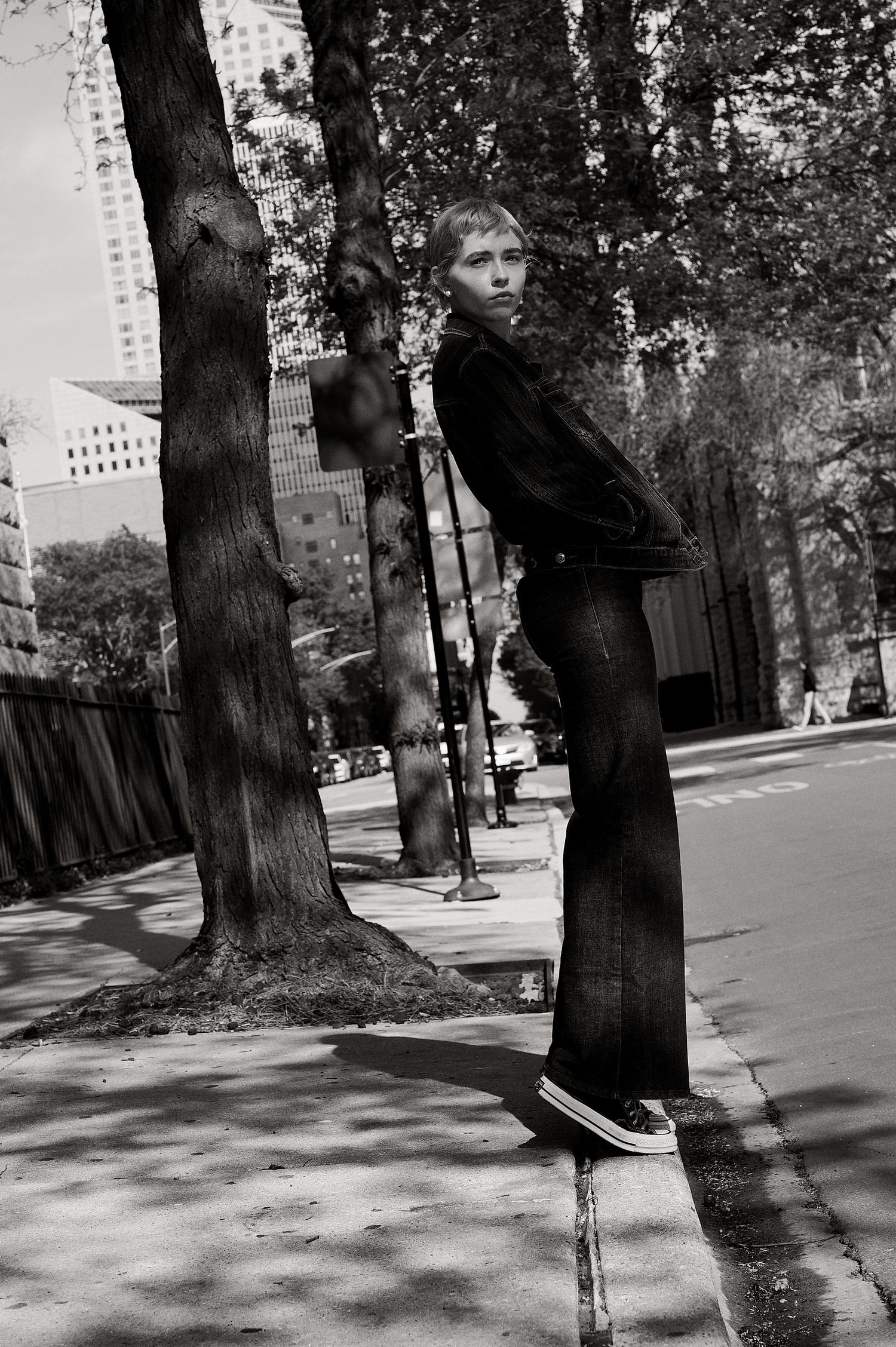Young woman in black flared jeans and Converse sneakers poses on urban sidewalk near trees in black and white photo.
