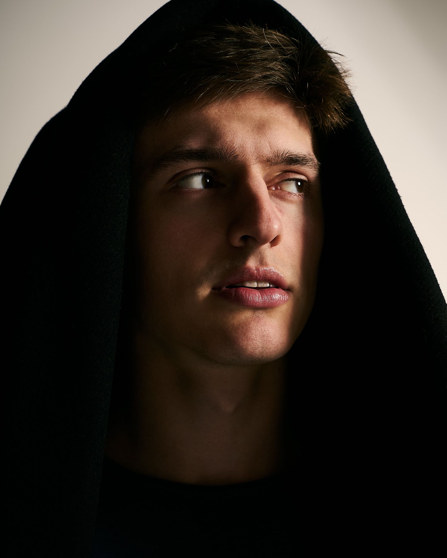 Dramatic portrait of a young man with a black hood framing his face against a light background.
