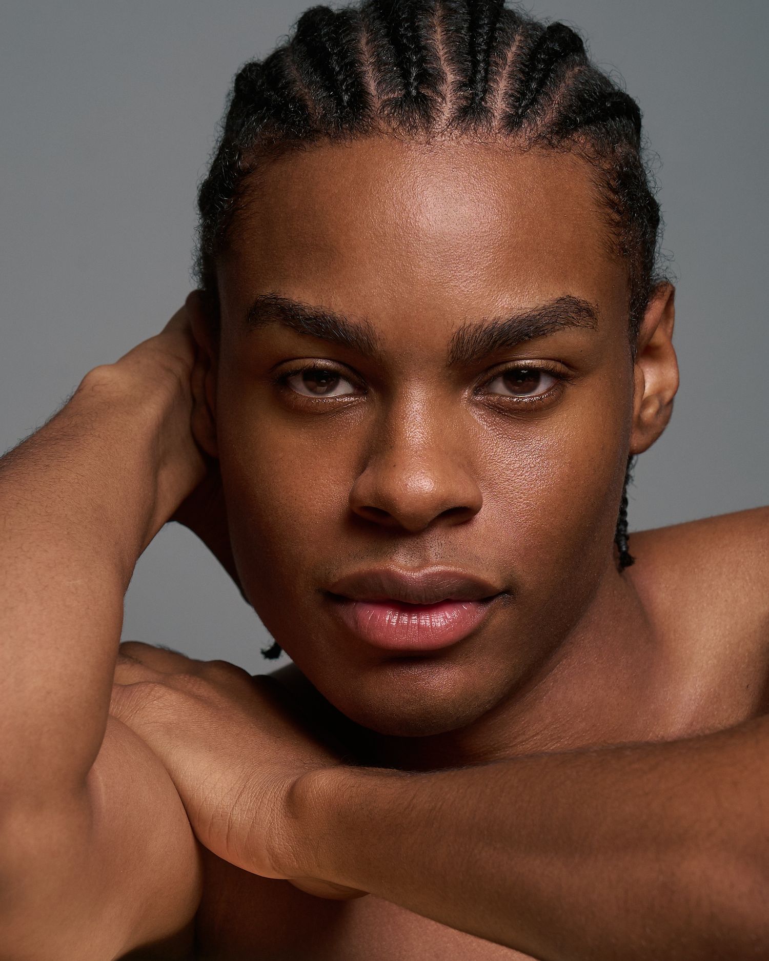 Close-up portrait of a male model with cornrow braids resting his chin on crossed arms.