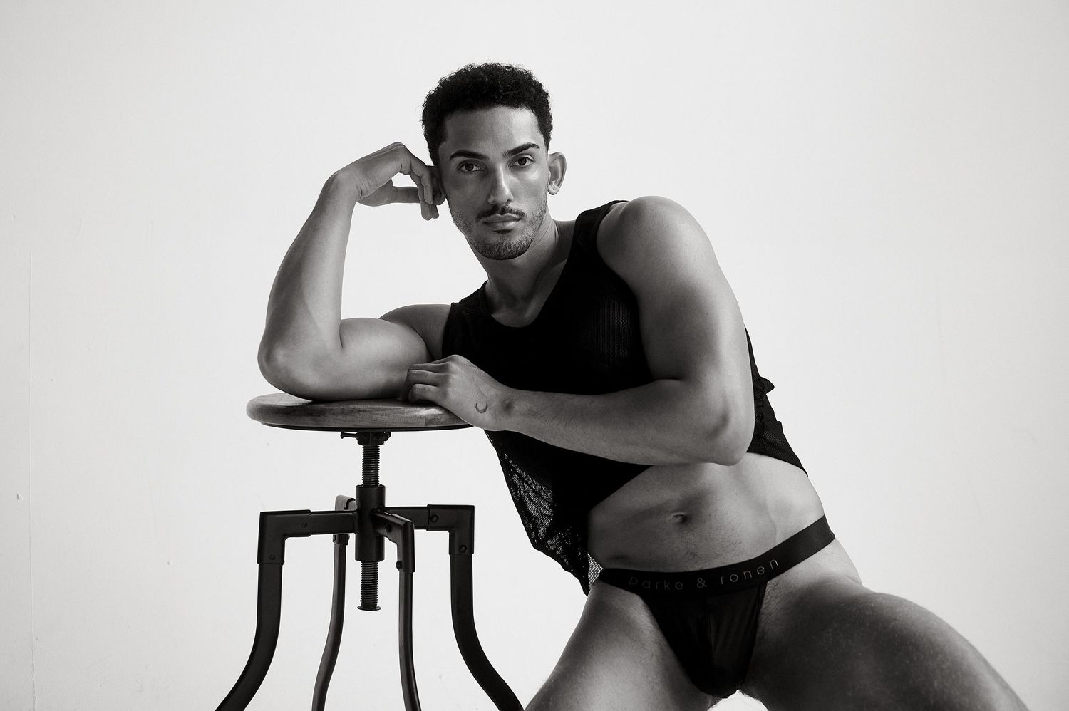 Muscular male model in black tank and briefs posing on a stool, flexing bicep in a black and white photo.