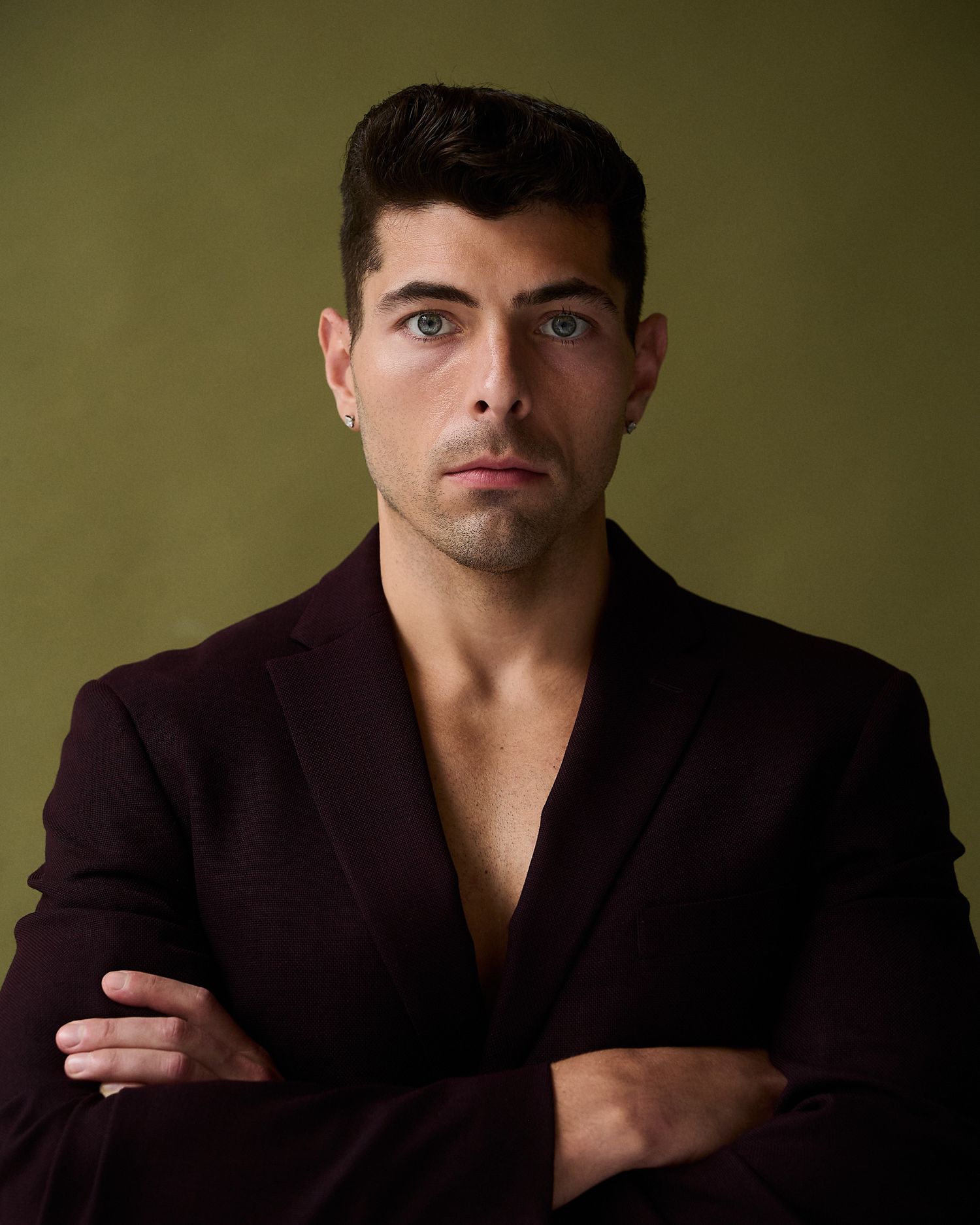 Man in dark burgundy blazer with arms crossed poses against olive green background.