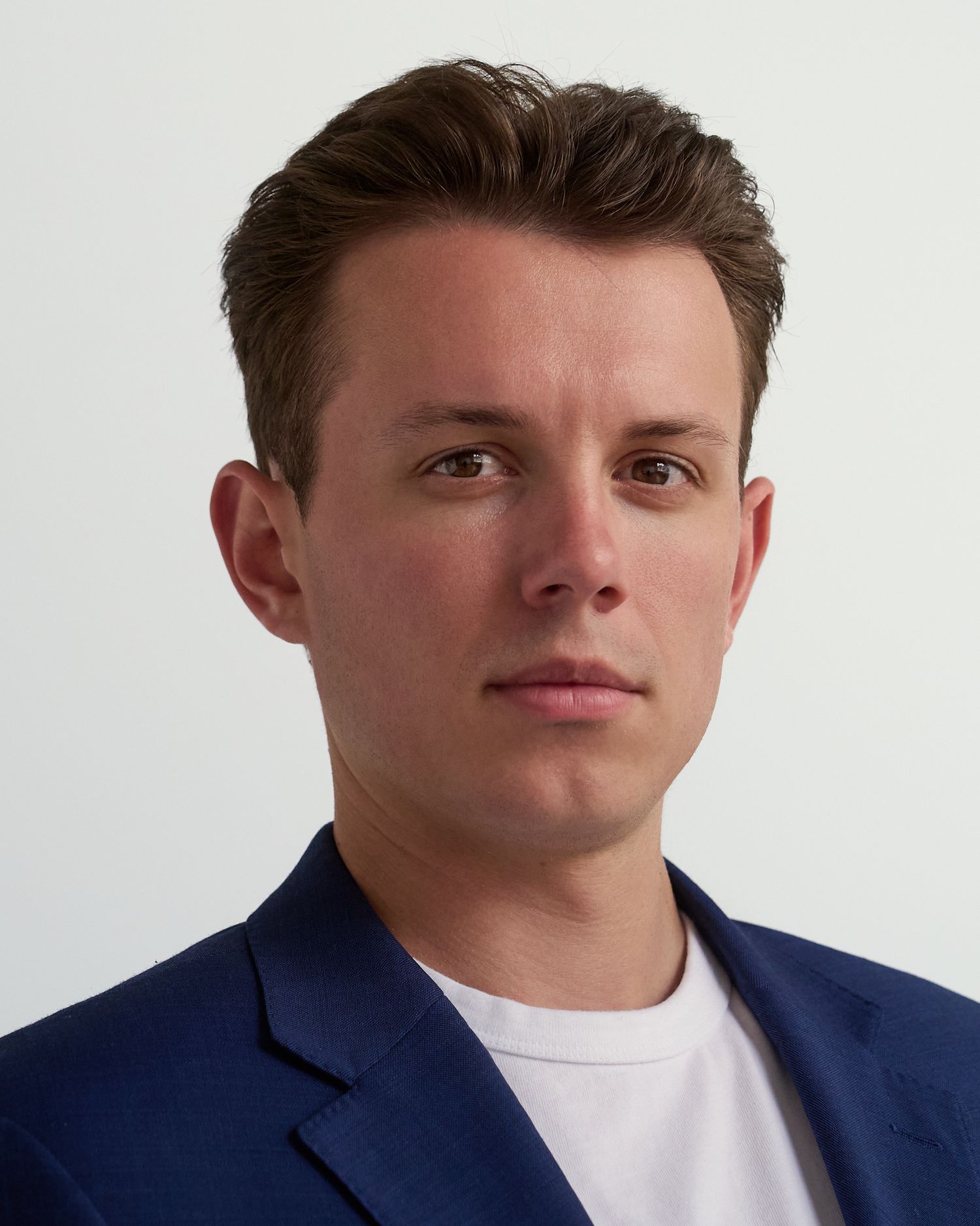 Young man in blue blazer and white t-shirt poses for professional headshot against white background.