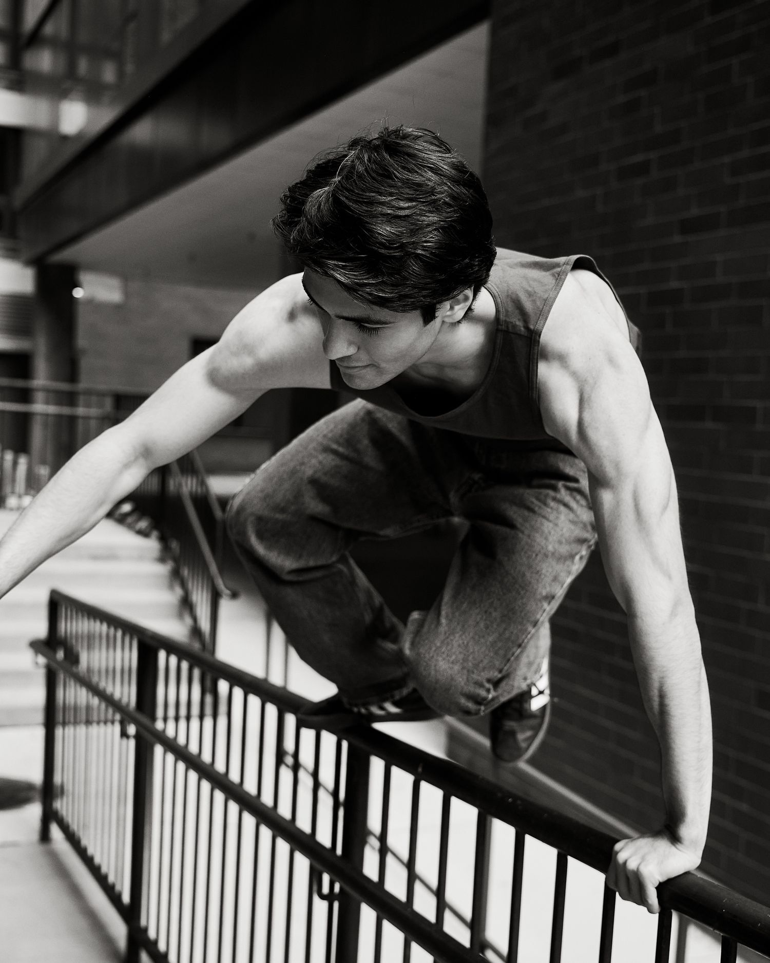 A young athletic man in a tank top and jeans balances on a railing in a black and white urban photo.