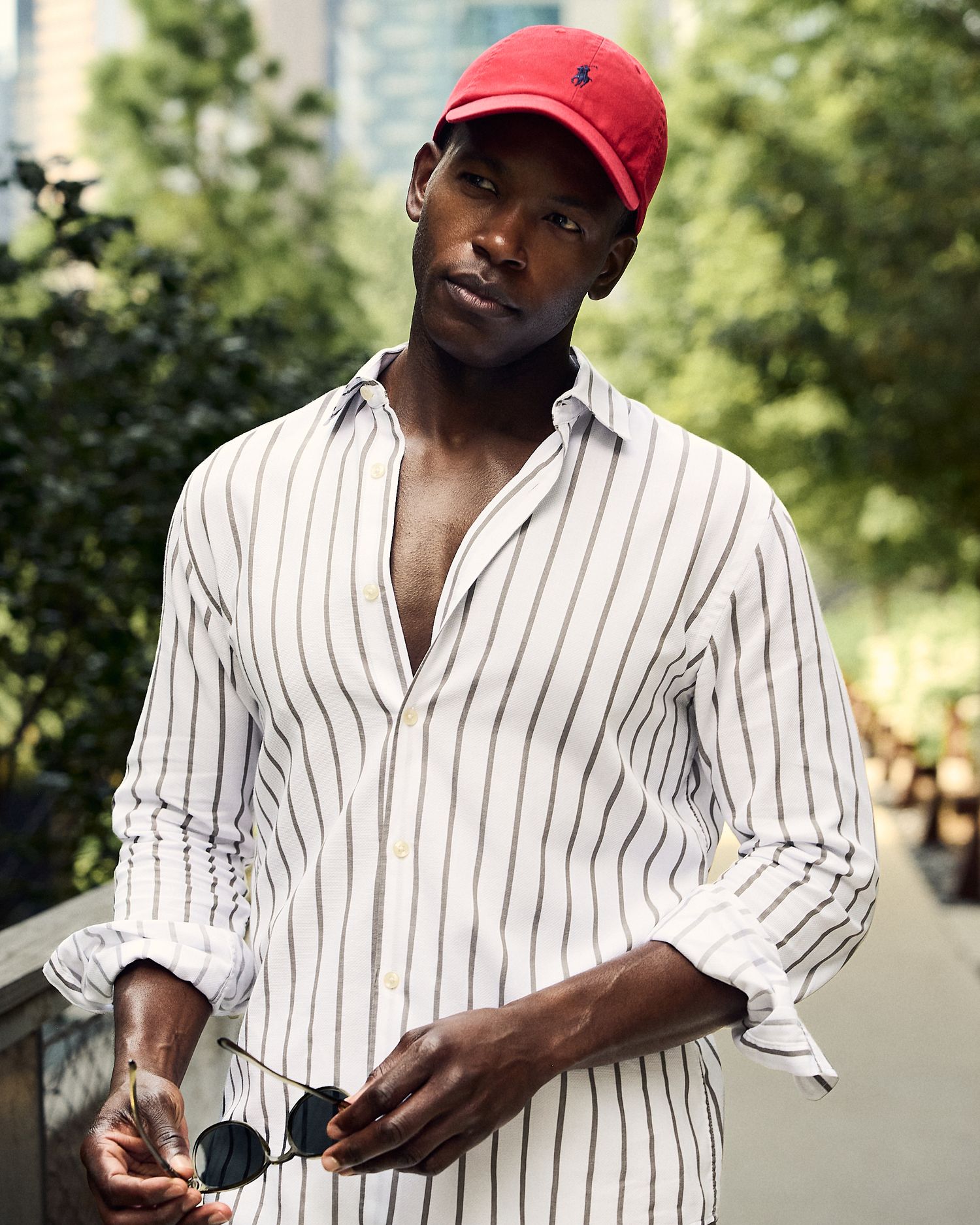 Man in red Polo Ralph Lauren cap and striped white shirt holding sunglasses outdoors.