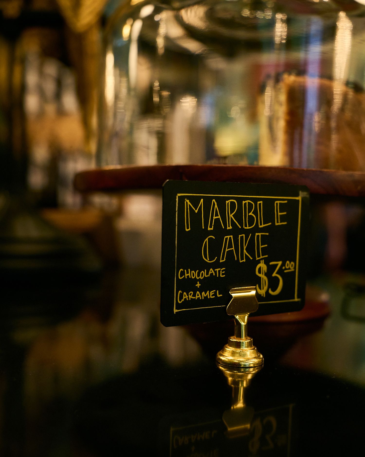 A chalkboard sign for marble cake with chocolate and caramel priced at $3.50 displayed under a glass dome.