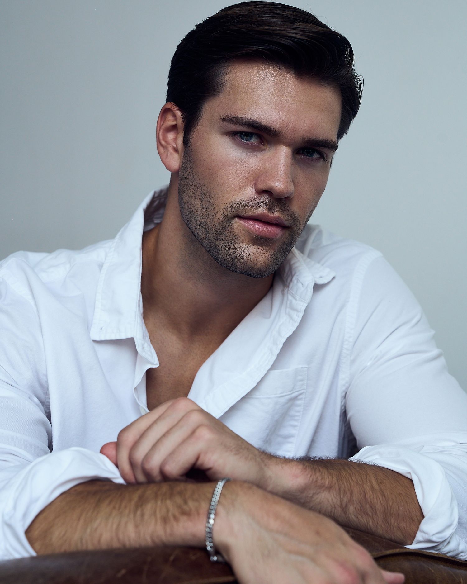 Young man in white shirt with silver bracelet poses confidently against neutral background.