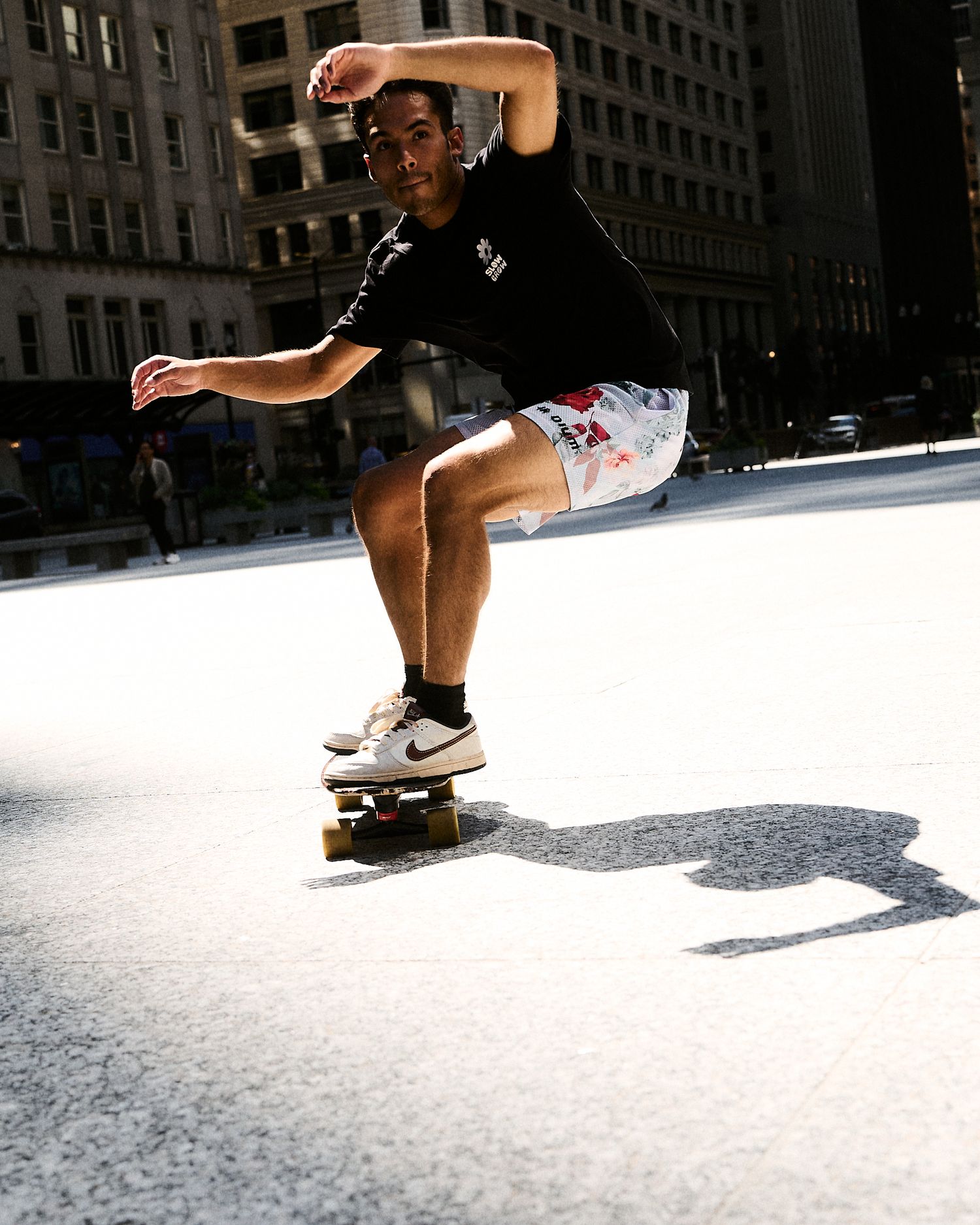 Young skateboarder performing a trick on a sunlit urban street wearing Nike sneakers and printed shorts.
