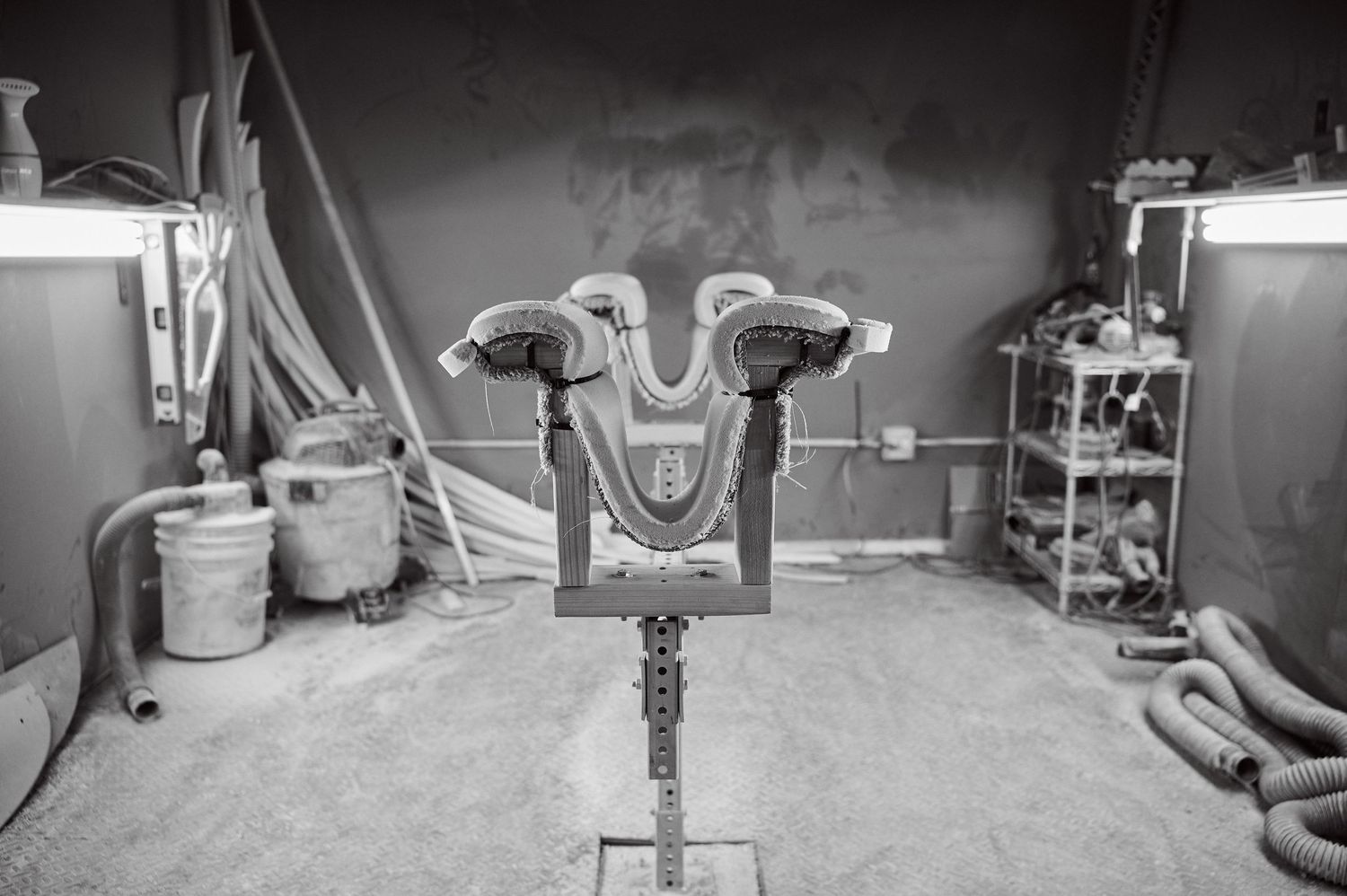 A surfboard shaping stand with fin boxes mounted in an industrial workshop under bright lights.