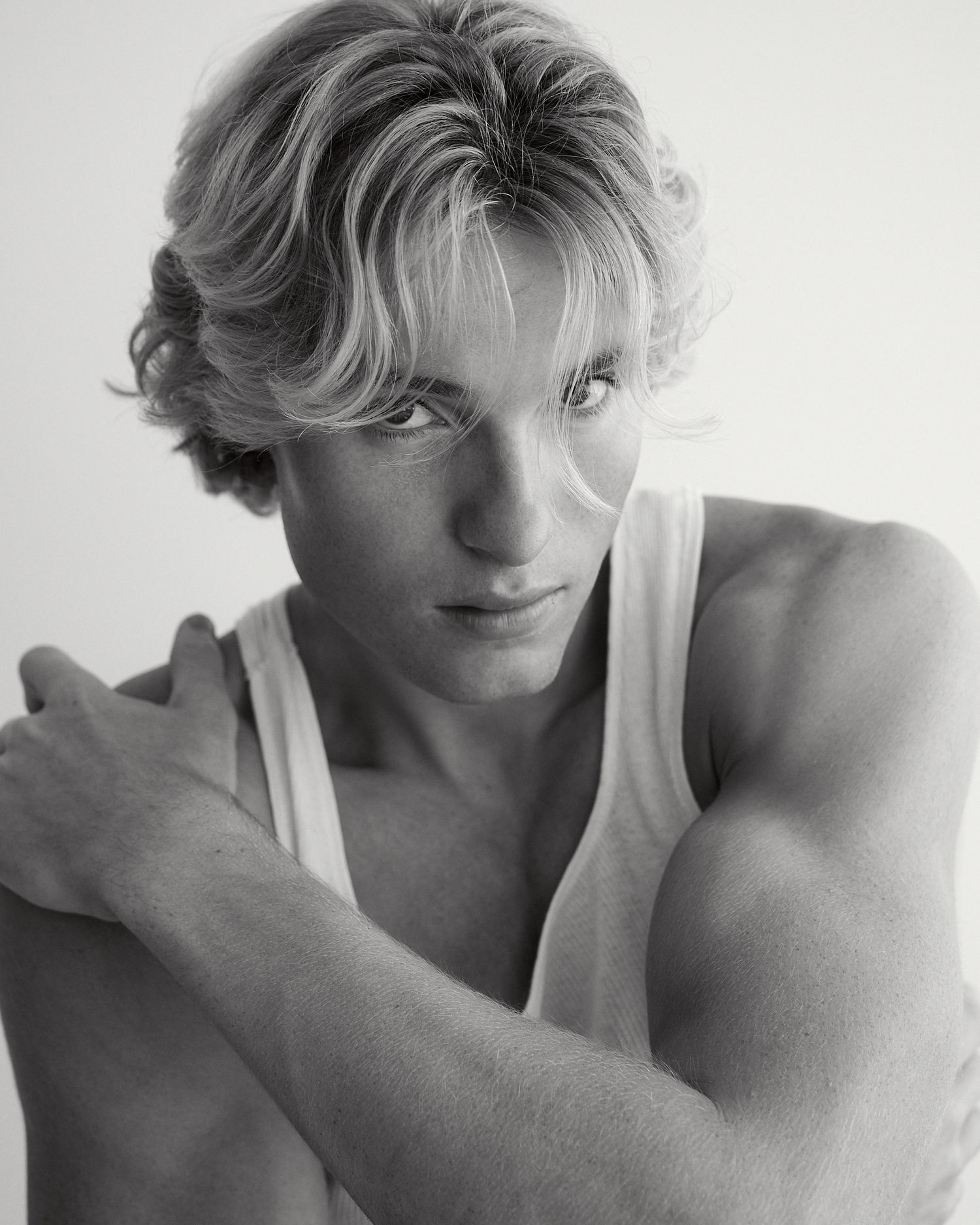 Black and white portrait of a young blonde male model in a white tank top posing with arms crossed.