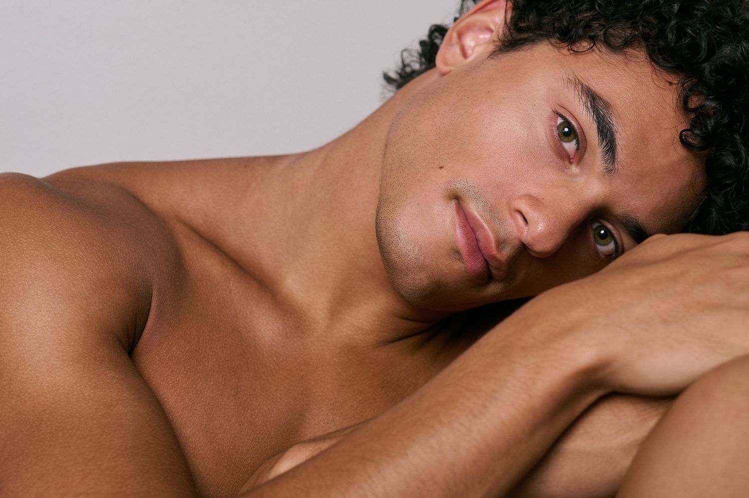 Close-up portrait of a shirtless young man with curly hair resting his head on his arms.