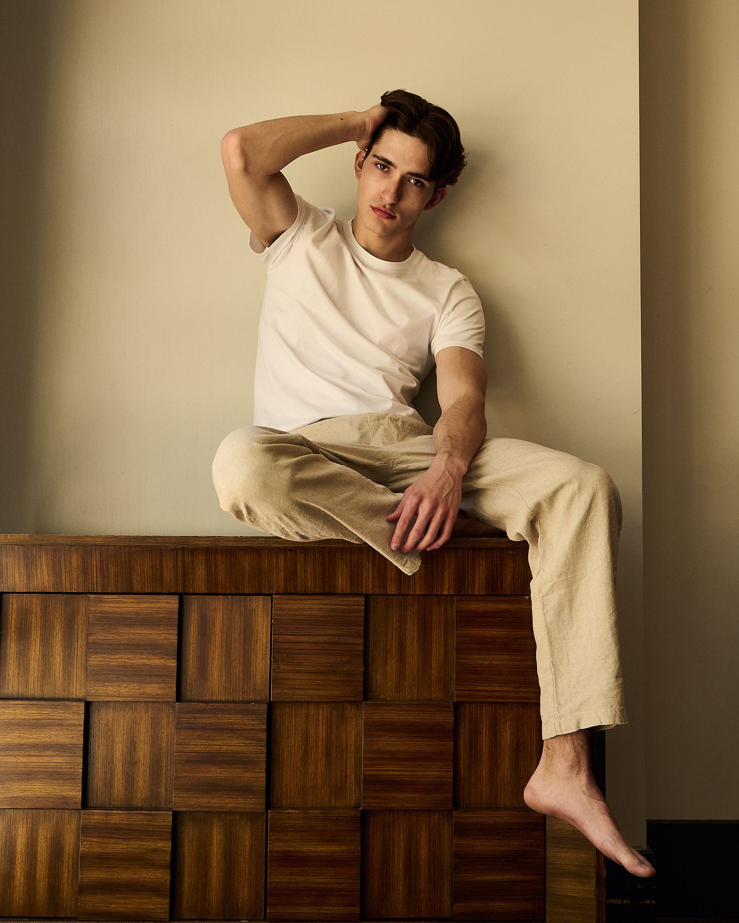 Young man in white tee and beige trousers sits barefoot atop a wooden dresser, posing with hand behind head.