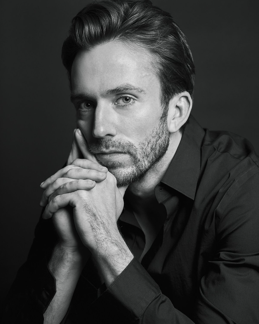 Professional black and white portrait of bearded man in dark shirt with hands clasped, studio lighting.